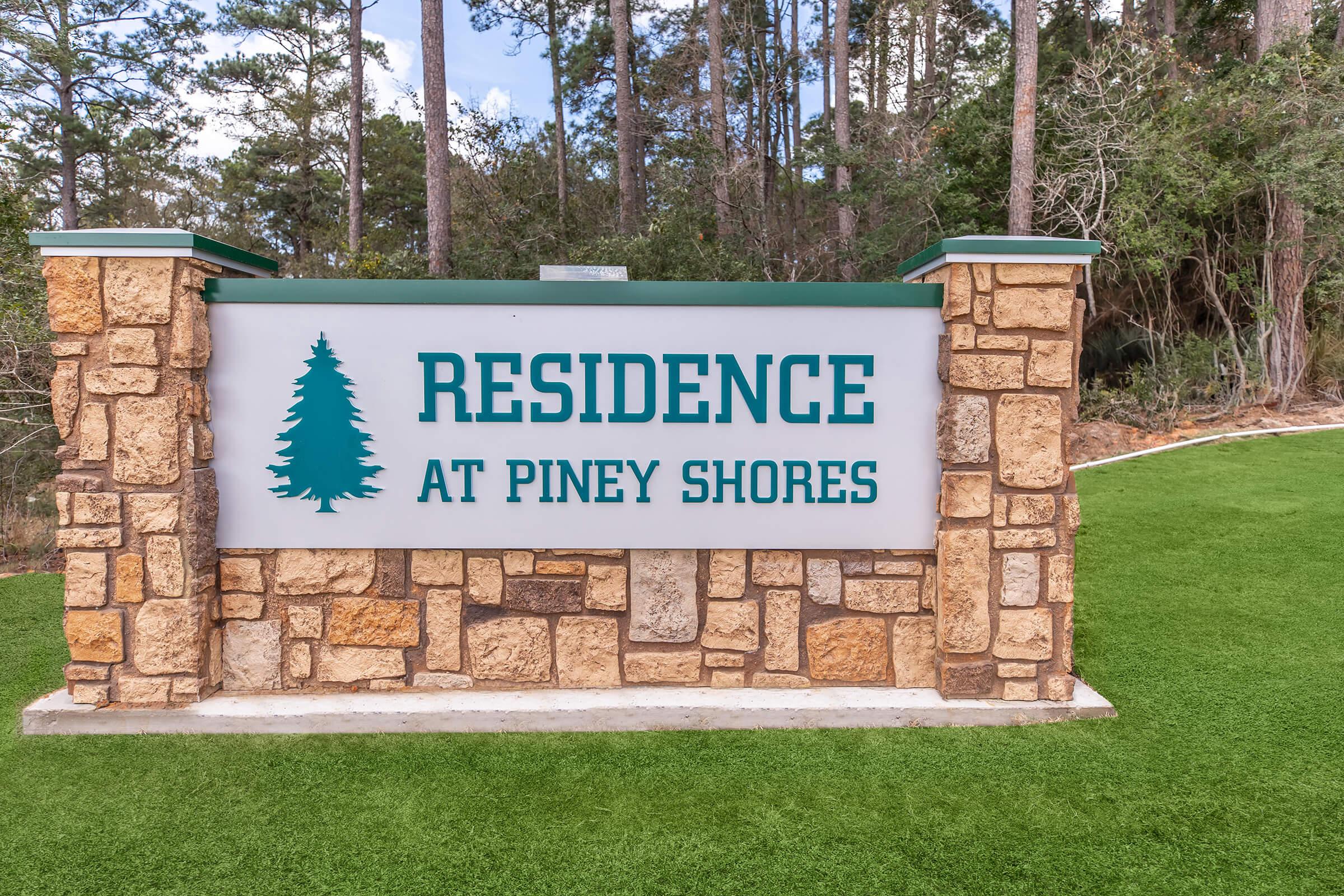 Sign for "Residence at Piney Shores" displayed prominently on a stone base, surrounded by green grass and trees in the background.