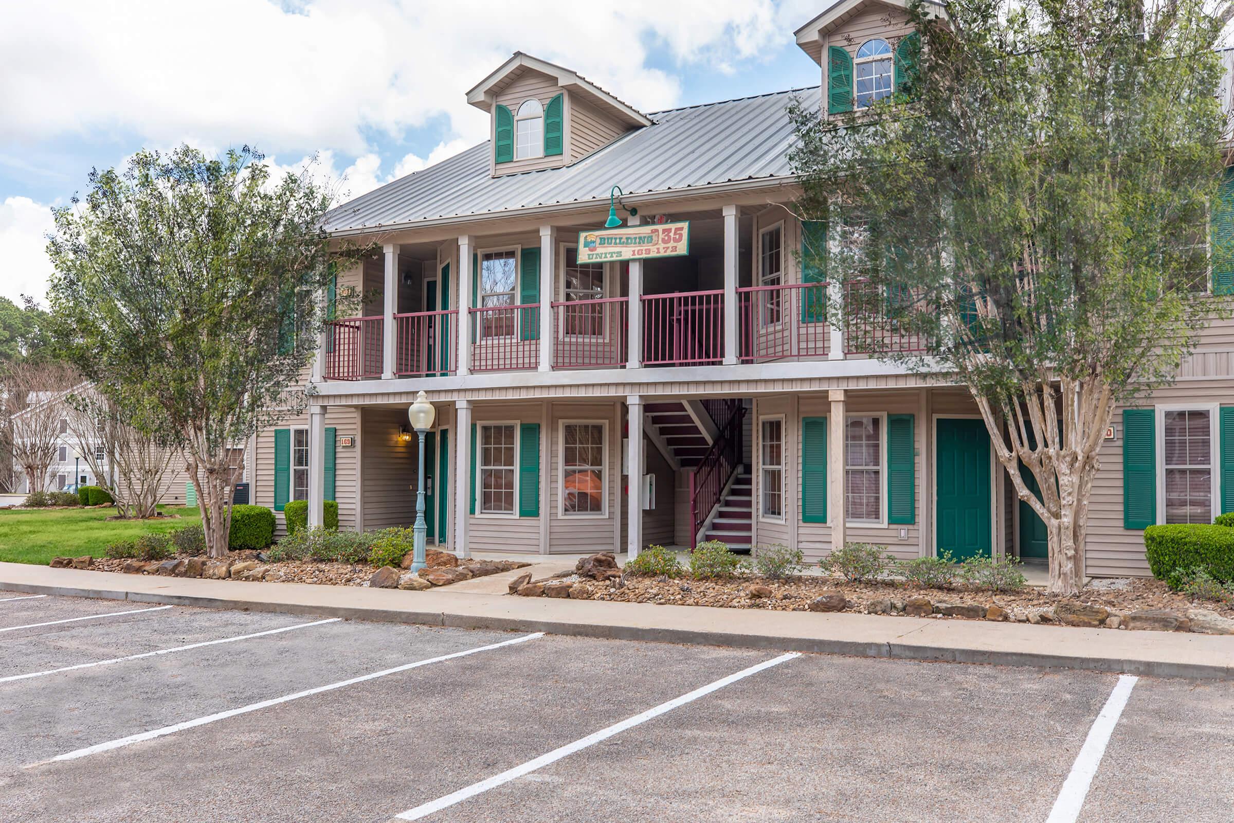 A two-story building with wooden siding and green shutters. It features a porch with railings, a staircase leading to the second floor, and several windows. The exterior is well-maintained, and there is a landscaped area with shrubs. A parking lot is visible in the foreground.