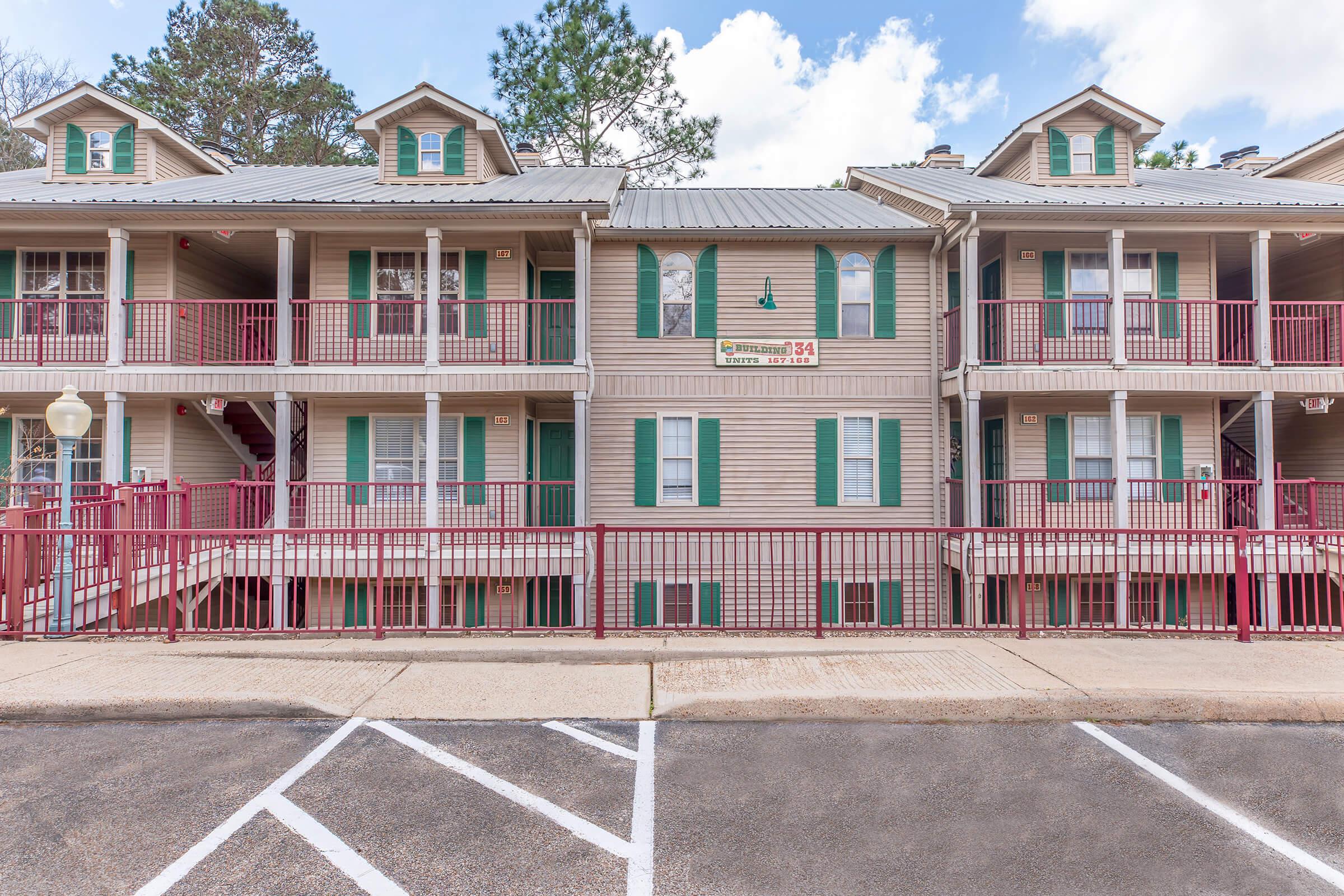 Exterior view of a multi-story residential building featuring a light-colored façade, green shutters, and a metal roof. The building has multiple balconies with red railings and is surrounded by a paved area with parking spaces. Trees and blue sky are visible in the background.