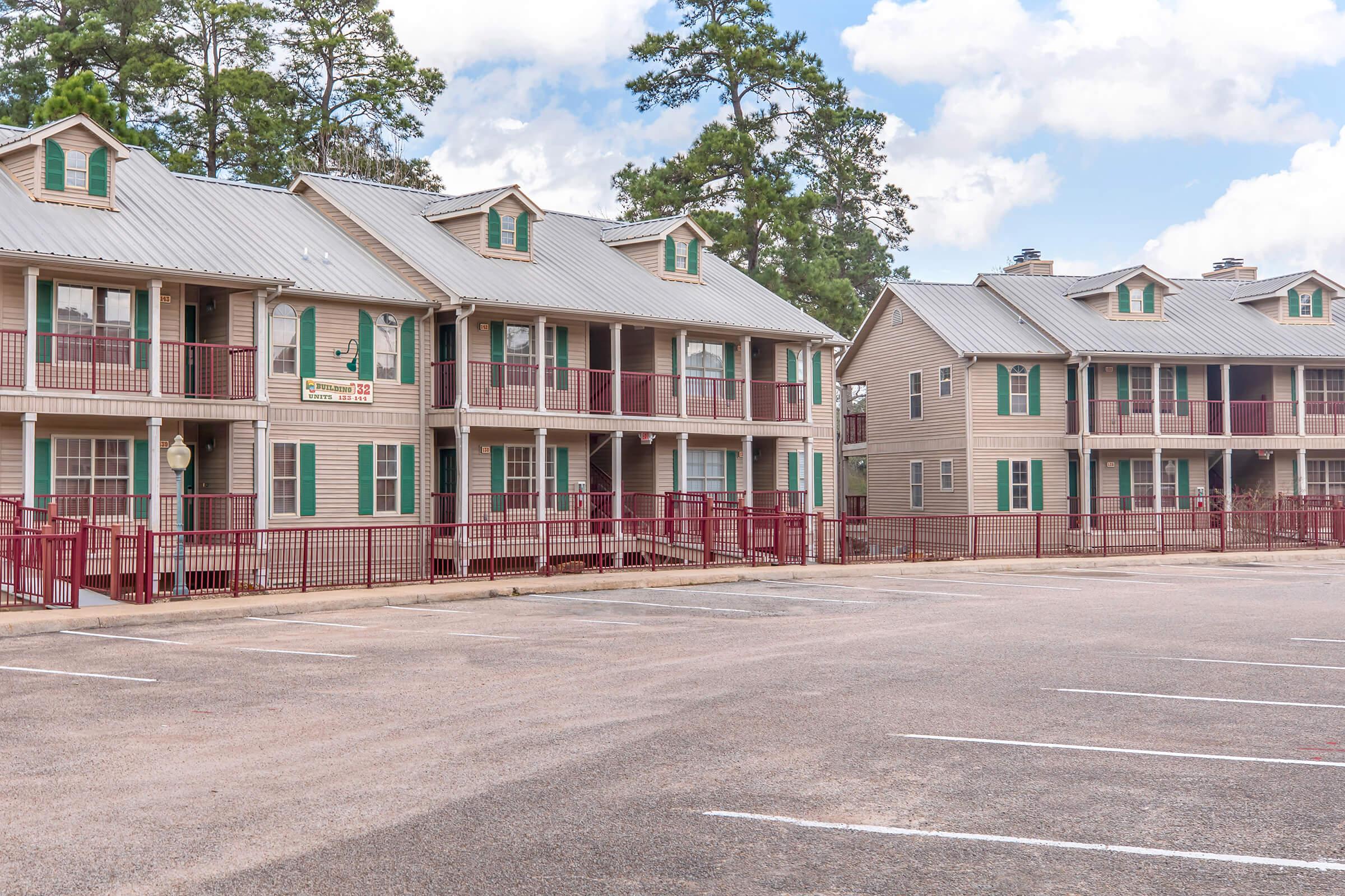 Two multi-unit residential buildings with metal roofs and green shutters, each featuring balconies and staircases. The buildings are situated in a spacious, paved area with empty parking spaces. Trees are visible in the background under a partly cloudy sky.