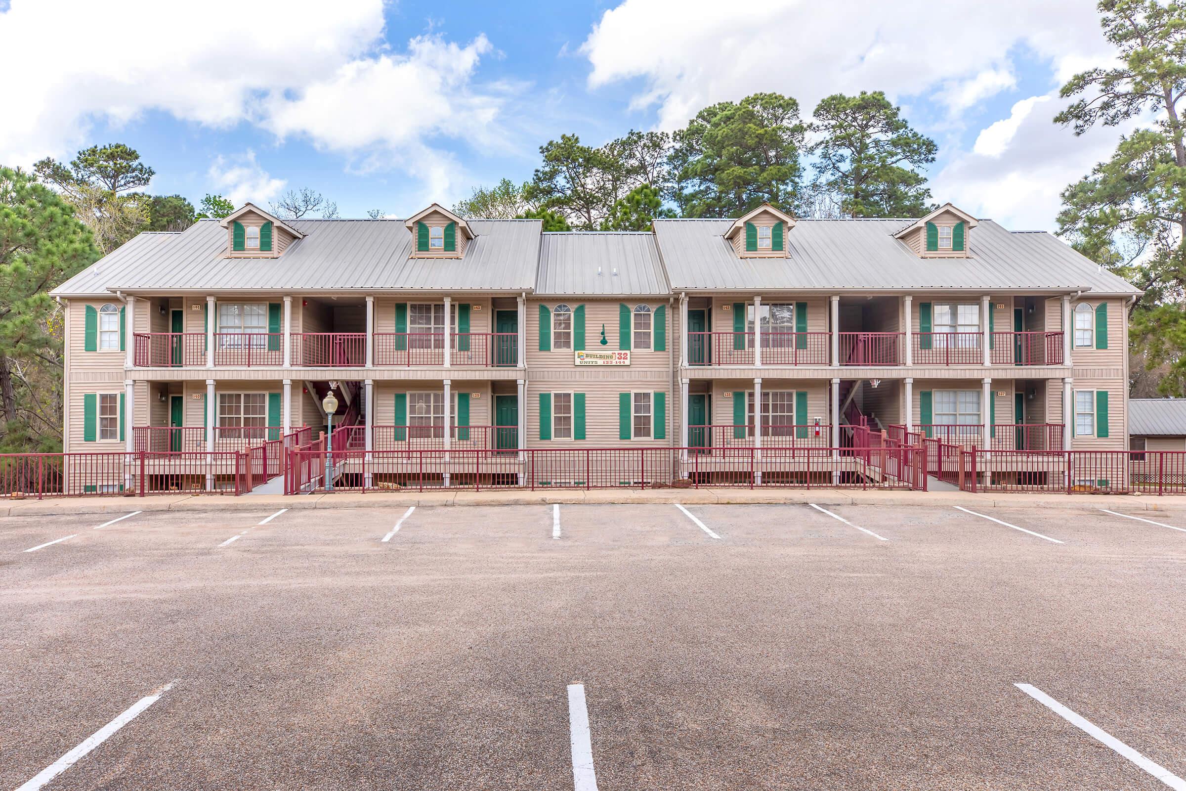 A two-story apartment building with several balconies, featuring a light-colored exterior and green shutters. The structure has a metal roof and is surrounded by trees. A parking lot is visible in the foreground, with multiple vacant parking spaces. The sky is partly cloudy.