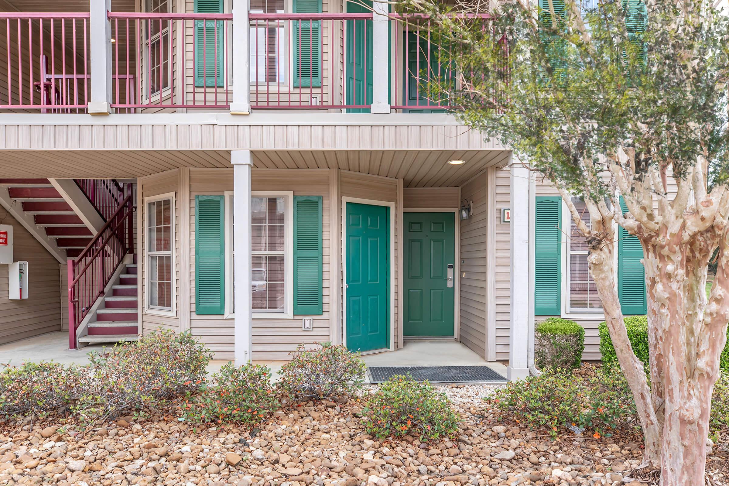 Exterior view of a residential building featuring green doors, several windows with shutters, and a small porch area. Surrounding the entrance are neatly maintained shrubs and decorative rocks. A staircase leads to the upper level, enhancing the multi-story structure's appeal.