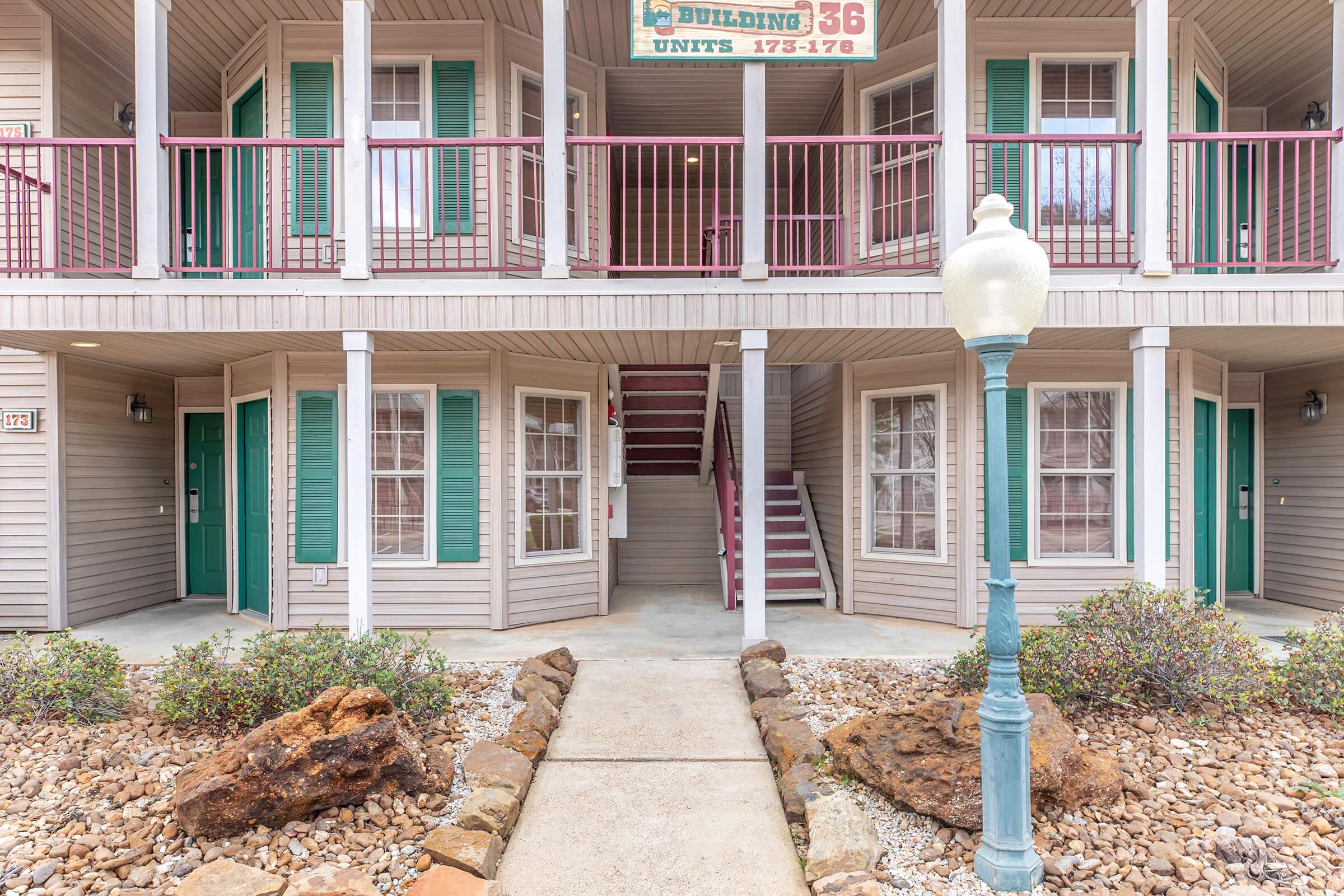 A multi-unit apartment building featuring two stories with light gray siding and green shutters. The entrance has several doors, and a decorative lamp post stands in front. Flower beds with rocks and plants line the pathway leading to the entrance, creating a welcoming atmosphere.