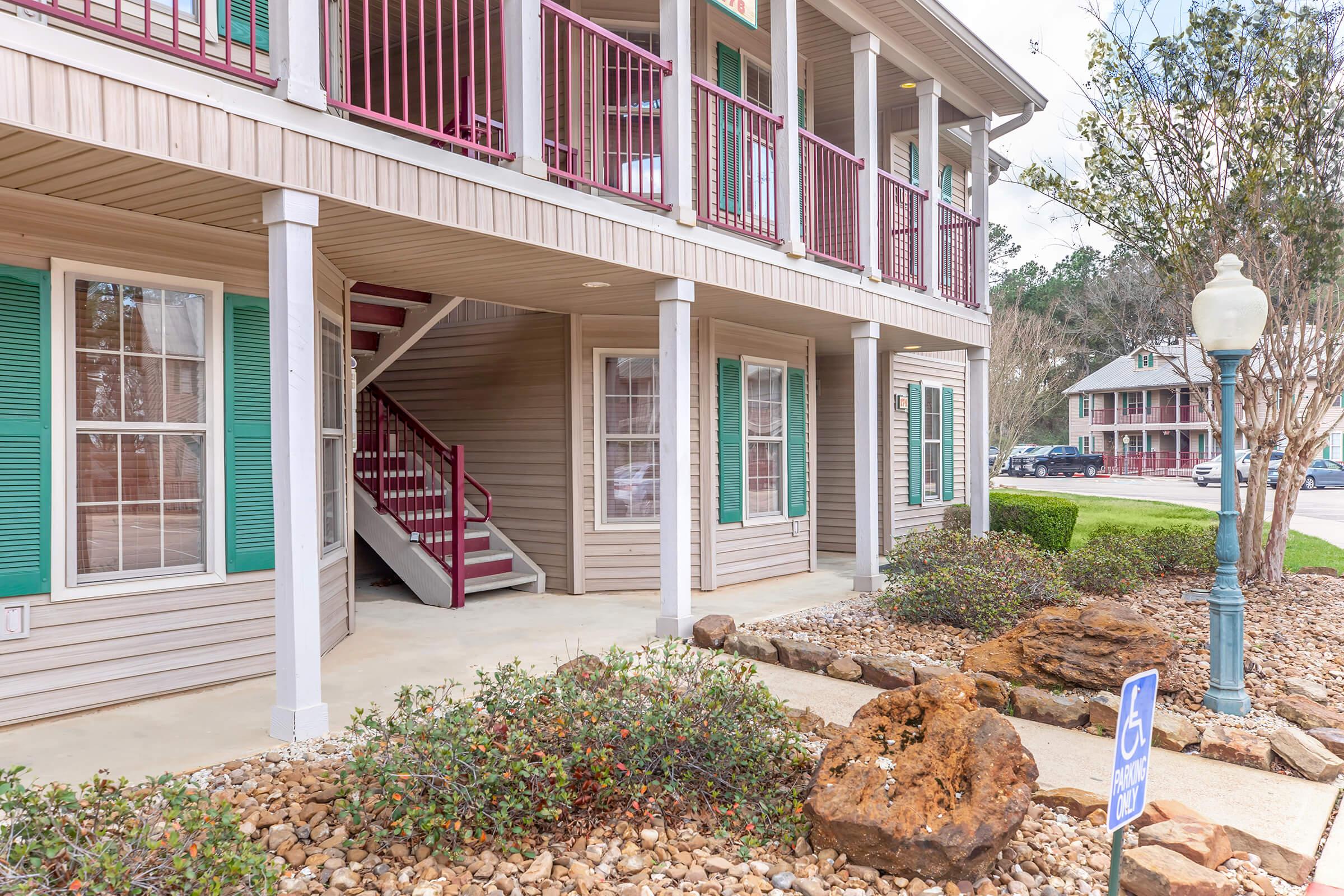Exterior view of a multi-story apartment building featuring covered porches with red railings. The ground level includes a set of outdoor stairs leading to upper floors. Surrounding the building are landscaped areas with rocks and vegetation, along with a lamp post and a designated accessible parking space nearby.