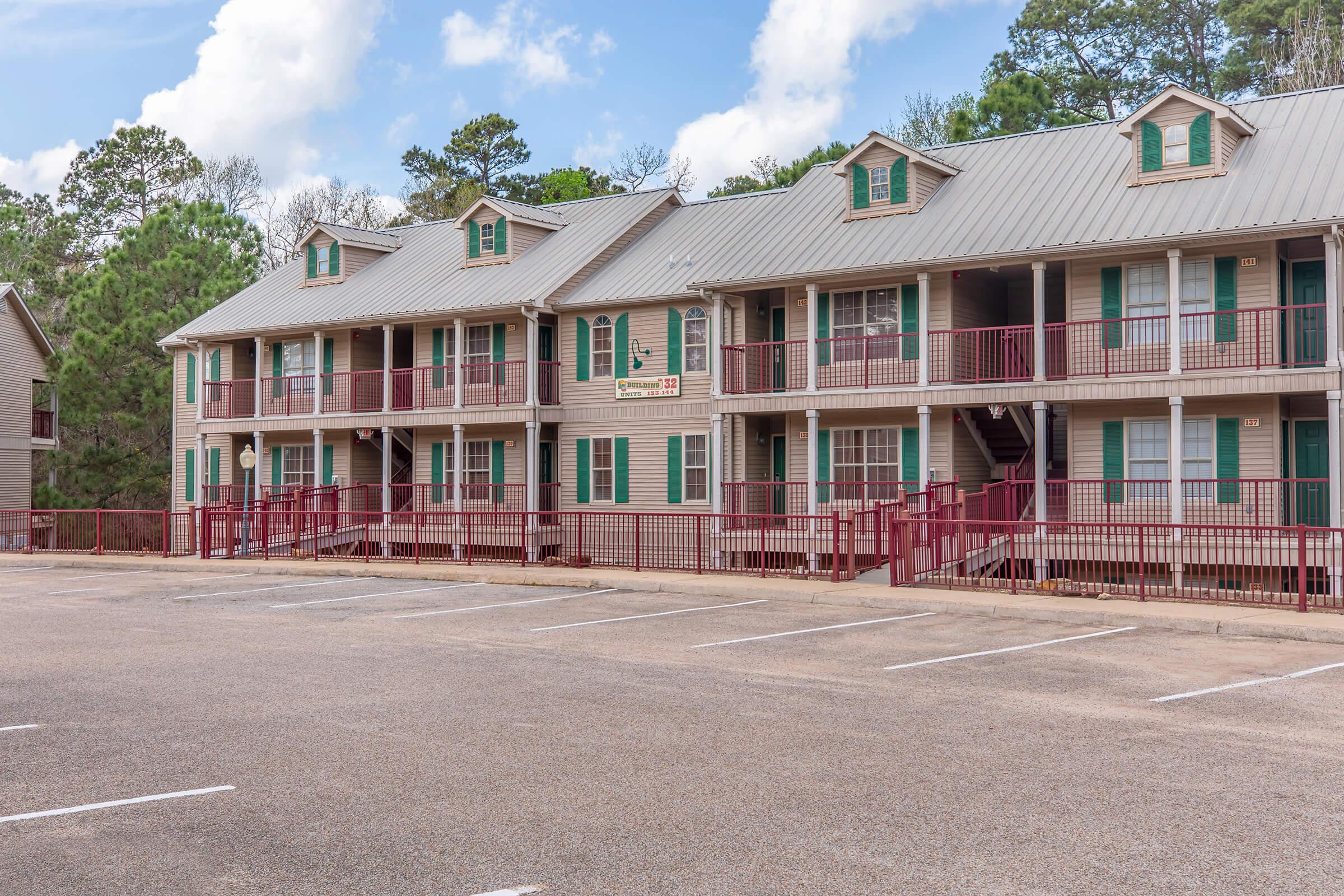 A two-story residential building with a red and beige color scheme. The building features multiple balconies with green shutters and a metal roof. A parking lot with several empty spaces is visible in front, along with trees in the background under a clear blue sky.