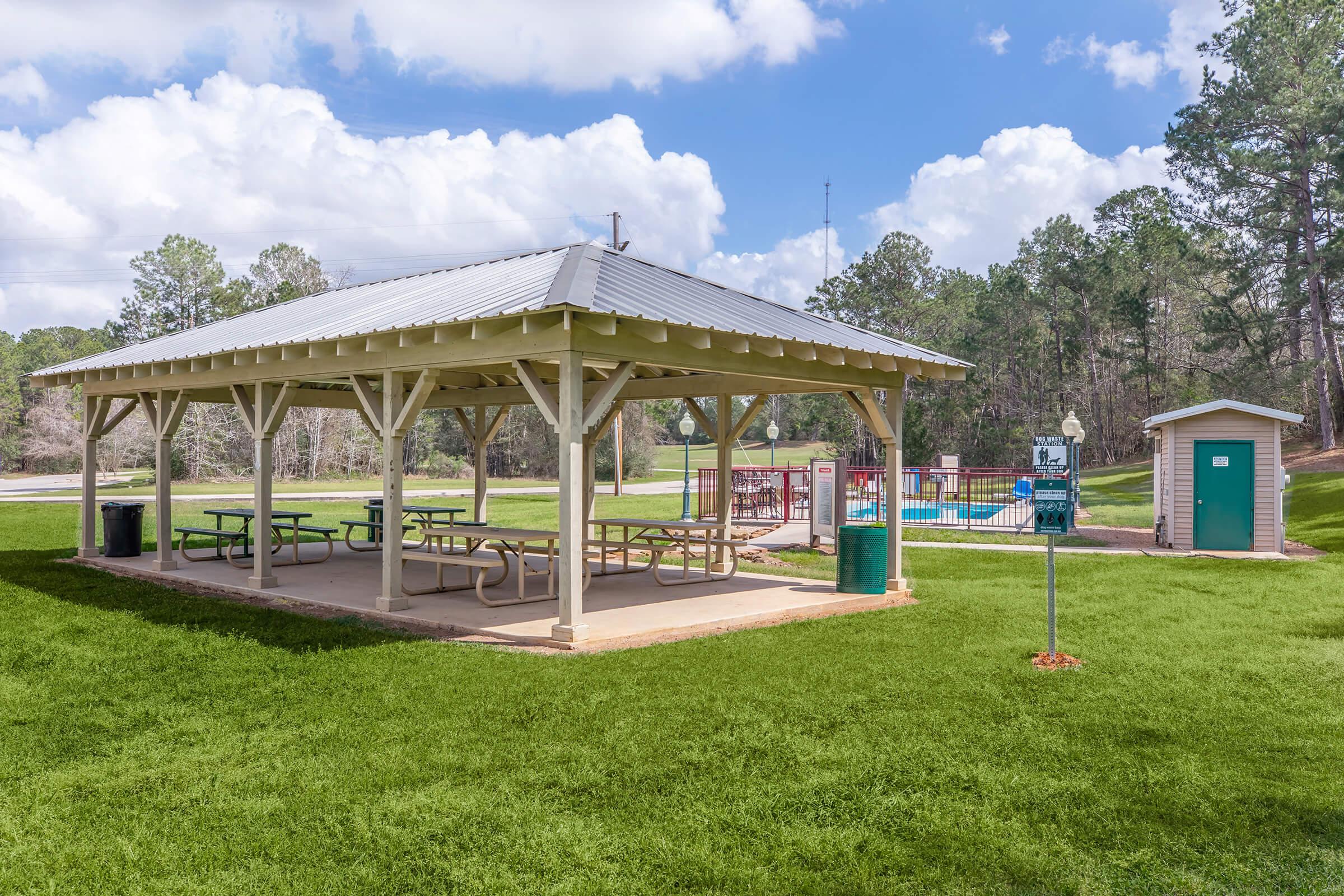 A covered picnic area with multiple tables sits on a well-maintained grassy space. In the background, there is a playground with colorful equipment and a small restroom building. The scene is set under a partly cloudy blue sky, surrounded by trees.