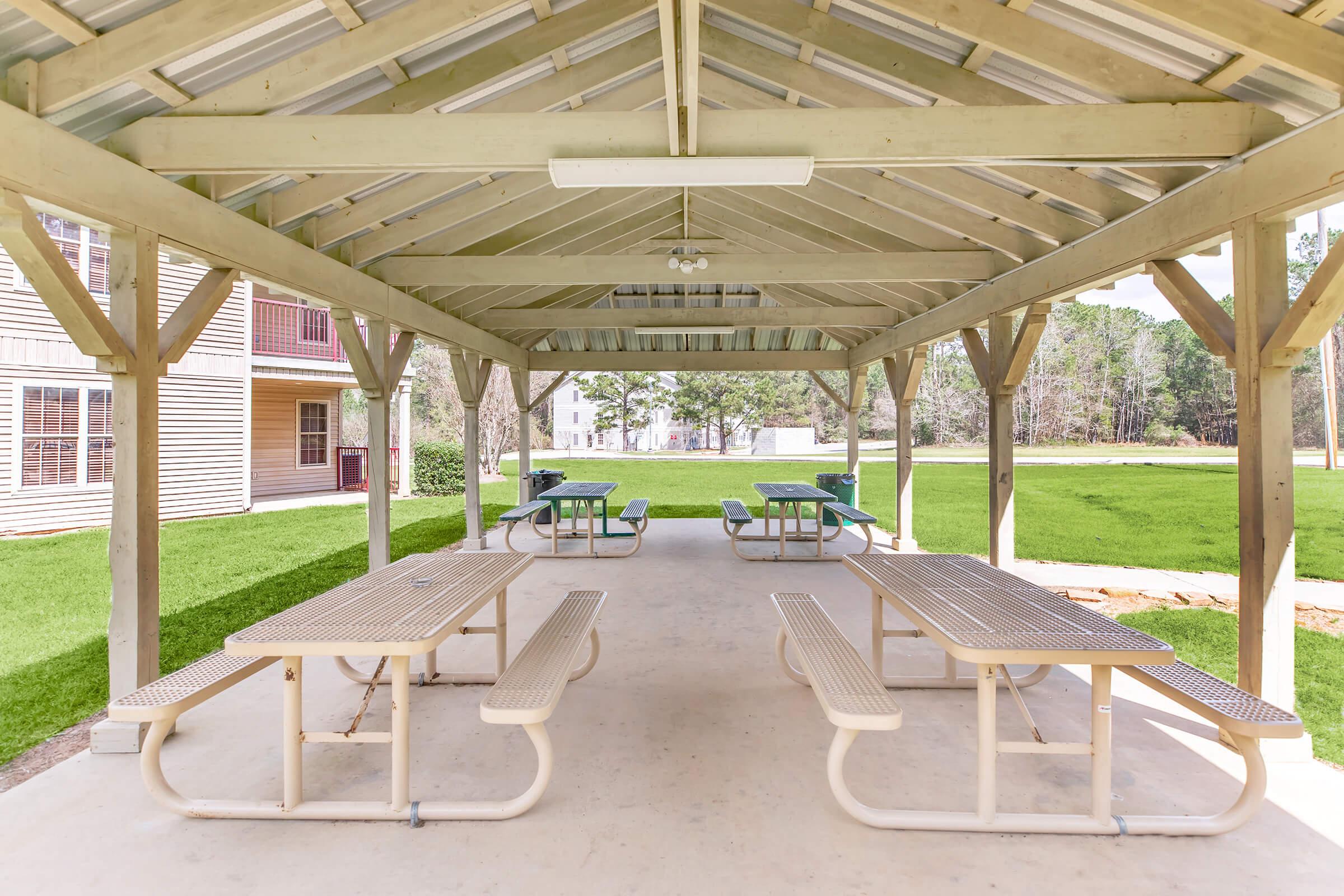 A covered outdoor picnic area with several tables and benches, surrounded by green grass and trees. The structure features a wooden framework and a metal roof, providing shade for visitors. There are multiple tables with light green tops, inviting for gatherings and meals.