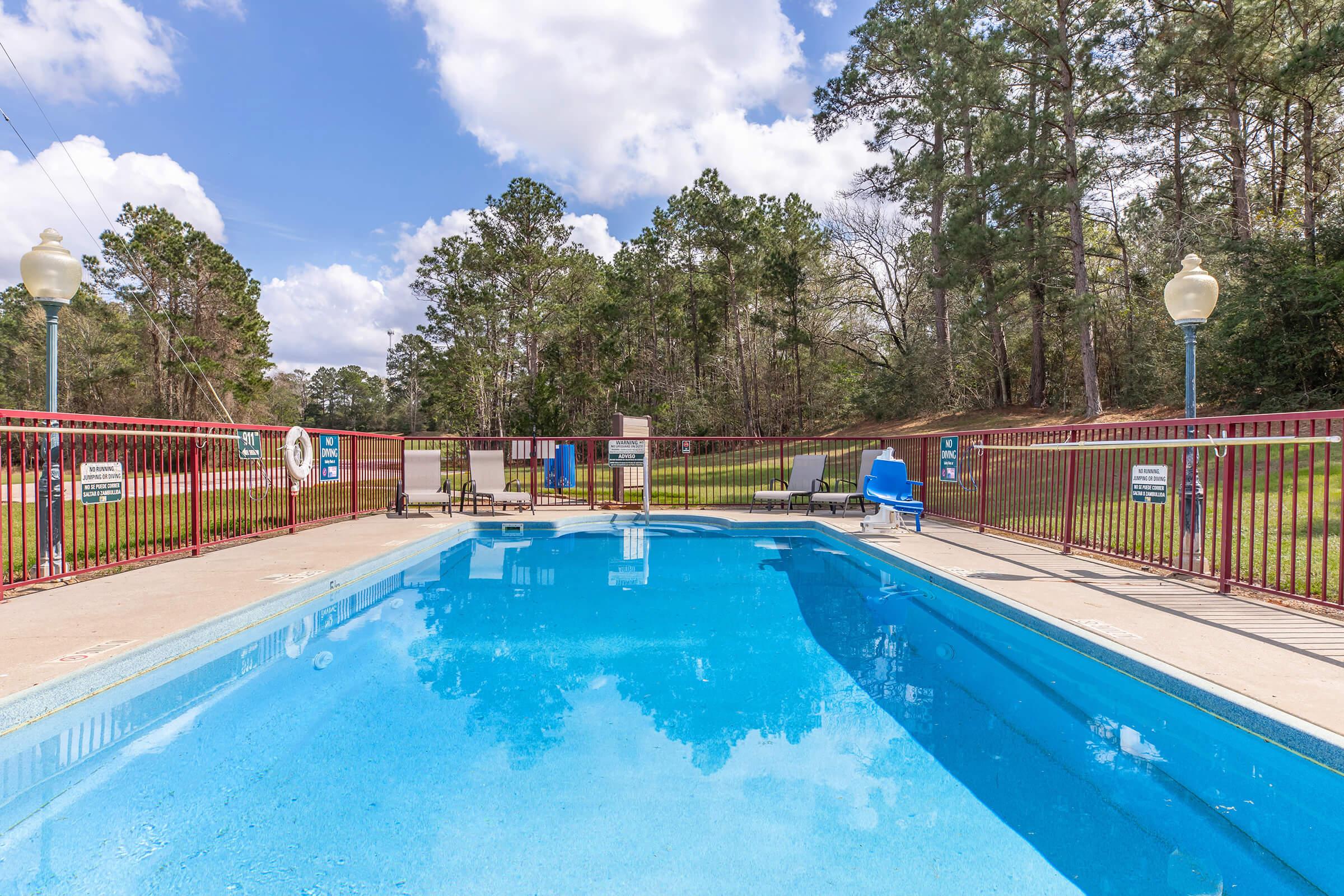 A clear blue swimming pool surrounded by a gated area. There are white lounge chairs placed around the pool, and green trees in the background under a partly cloudy sky. The scene conveys a relaxing outdoor atmosphere.