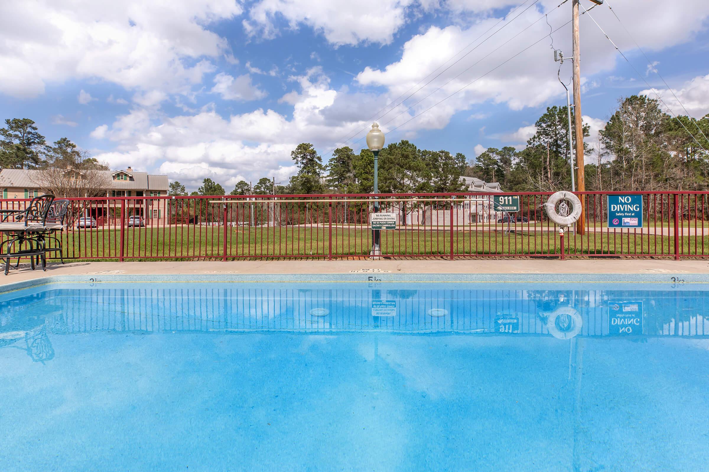 A clear swimming pool with a bright blue surface, surrounded by a red railing. In the background, there are grassy areas and buildings under a partly cloudy sky. There are signs visible near the pool, including a "No Diving" sign and a marker indicating emergency contact (911).