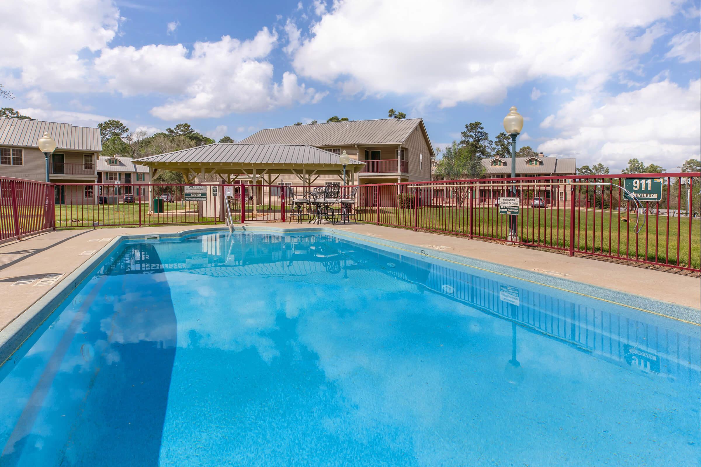 A clear blue swimming pool surrounded by a fenced area, with a gazebo nearby. Behind the pool are several multi-story residential buildings, set against a backdrop of a partly cloudy sky and green grass. The scene is bright and inviting, suggesting a recreational area for residents.