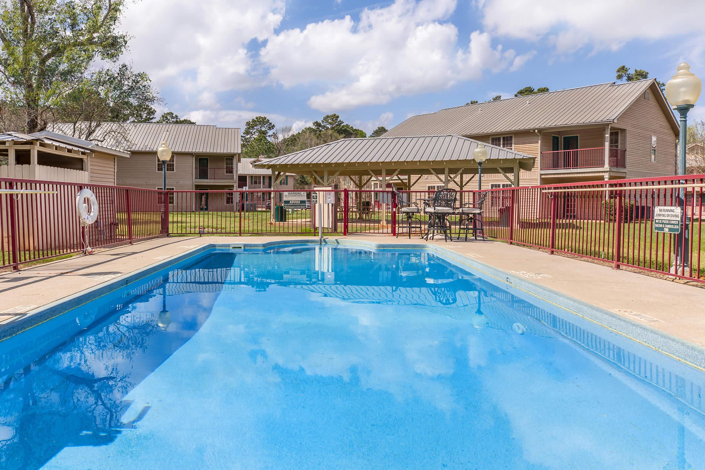 A clear blue swimming pool surrounded by a patio area with lounge chairs. In the background, there are two multi-story buildings with gray roofs and a green lawn. A shaded gazebo with picnic tables is visible near the pool. The sky is partially cloudy, giving a bright and inviting atmosphere.