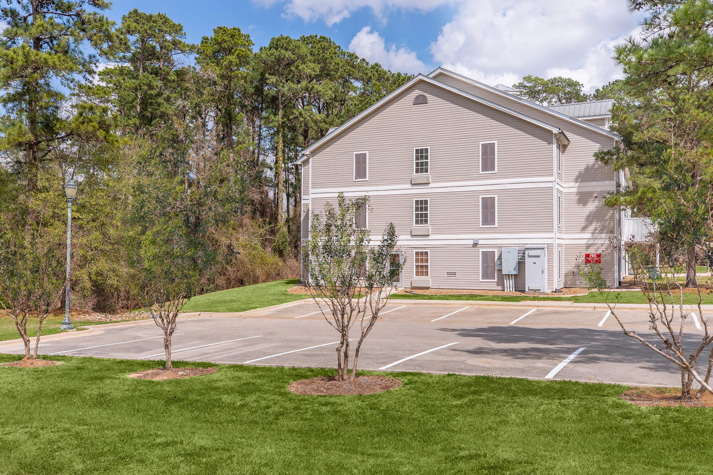 A two-story beige building with multiple windows surrounded by trees. In the foreground, a well-maintained green lawn with small trees and a parking area with marked spaces. The sky is partly cloudy.