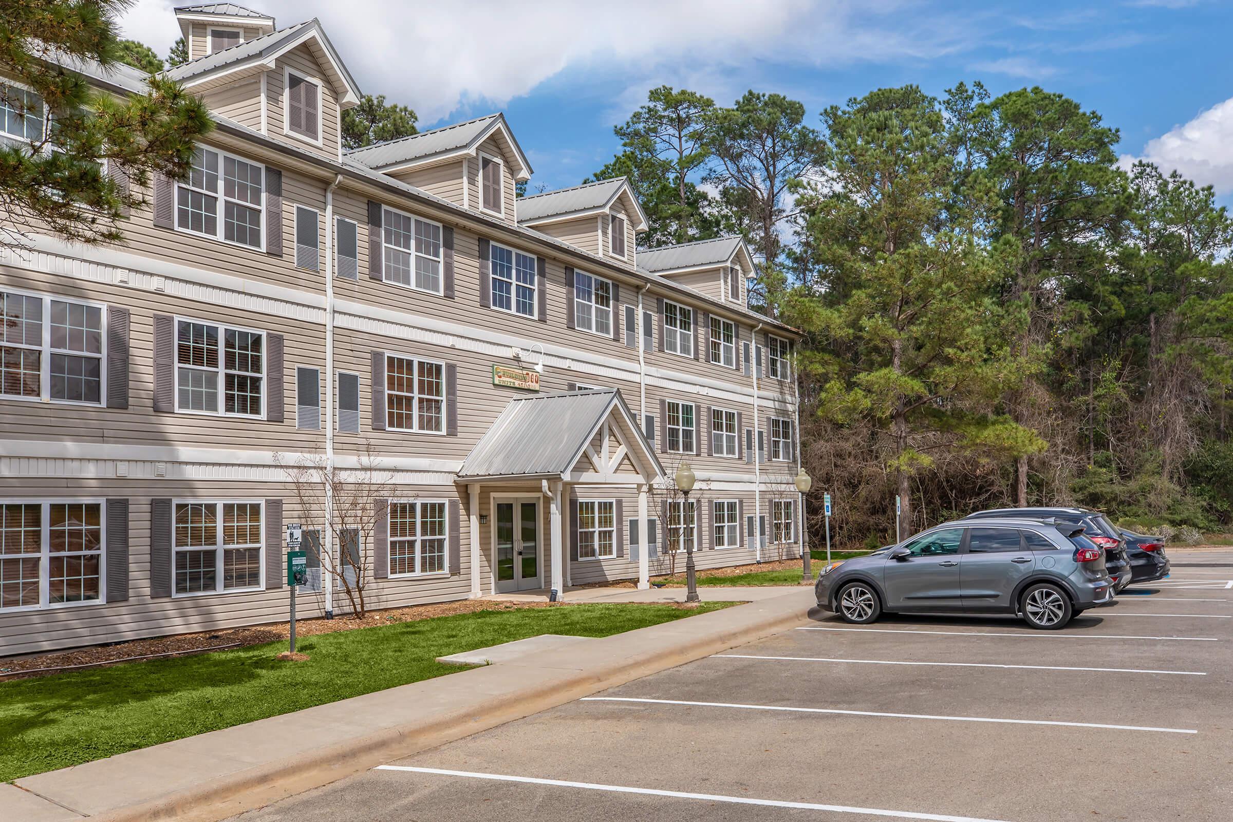A multi-story building with a light gray exterior and multiple windows. The front features a peaked roof and a welcoming entrance. A parking lot in front has several cars parked, and there are trees in the background, indicating a natural setting. The sky is partly cloudy.