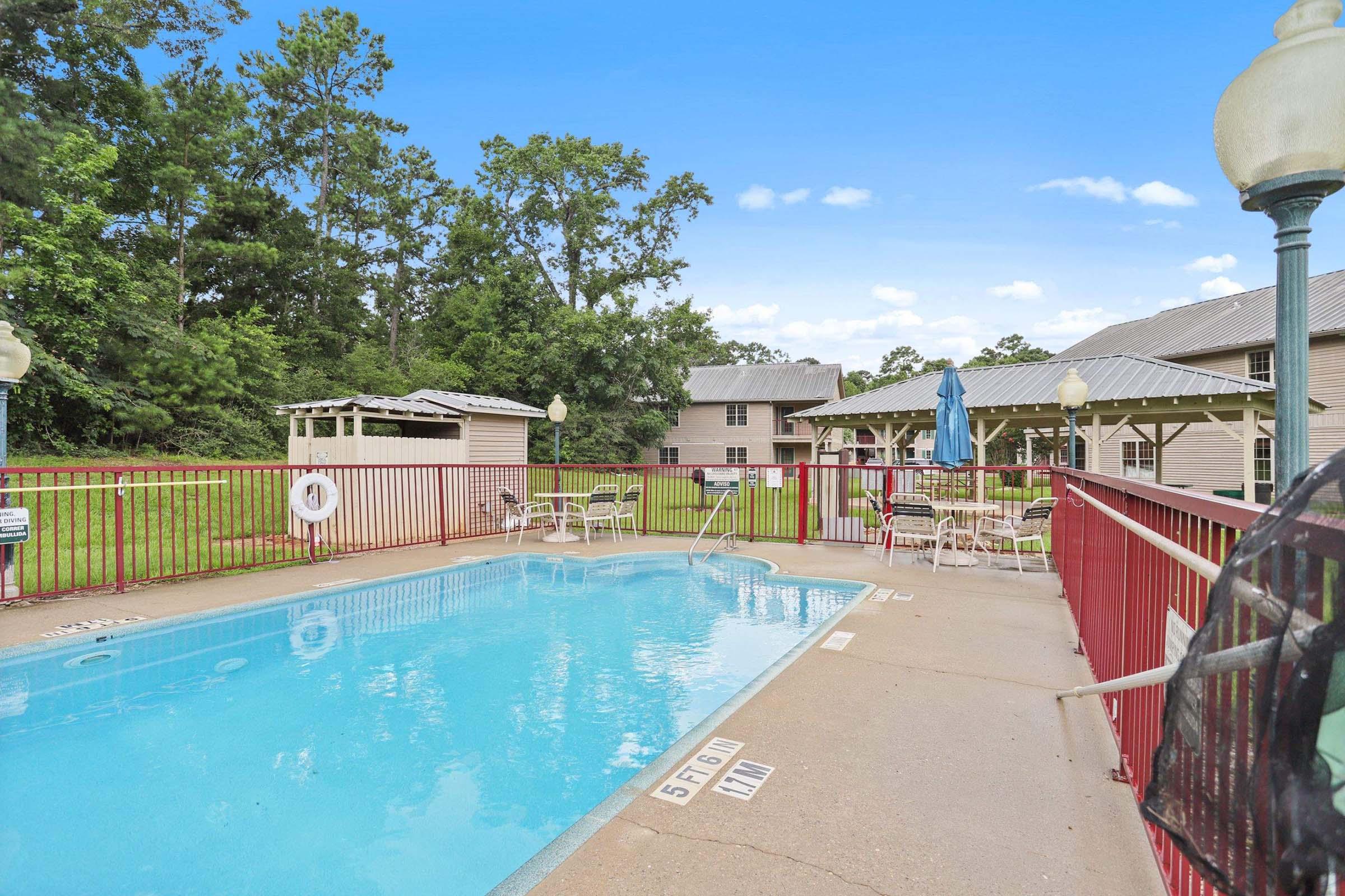 A clear swimming pool surrounded by a red railing. There are several chairs and tables nearby, with a blue umbrella. In the background, there are trees and a gazebo. The sky is bright blue with a few clouds, and a building is visible further back.