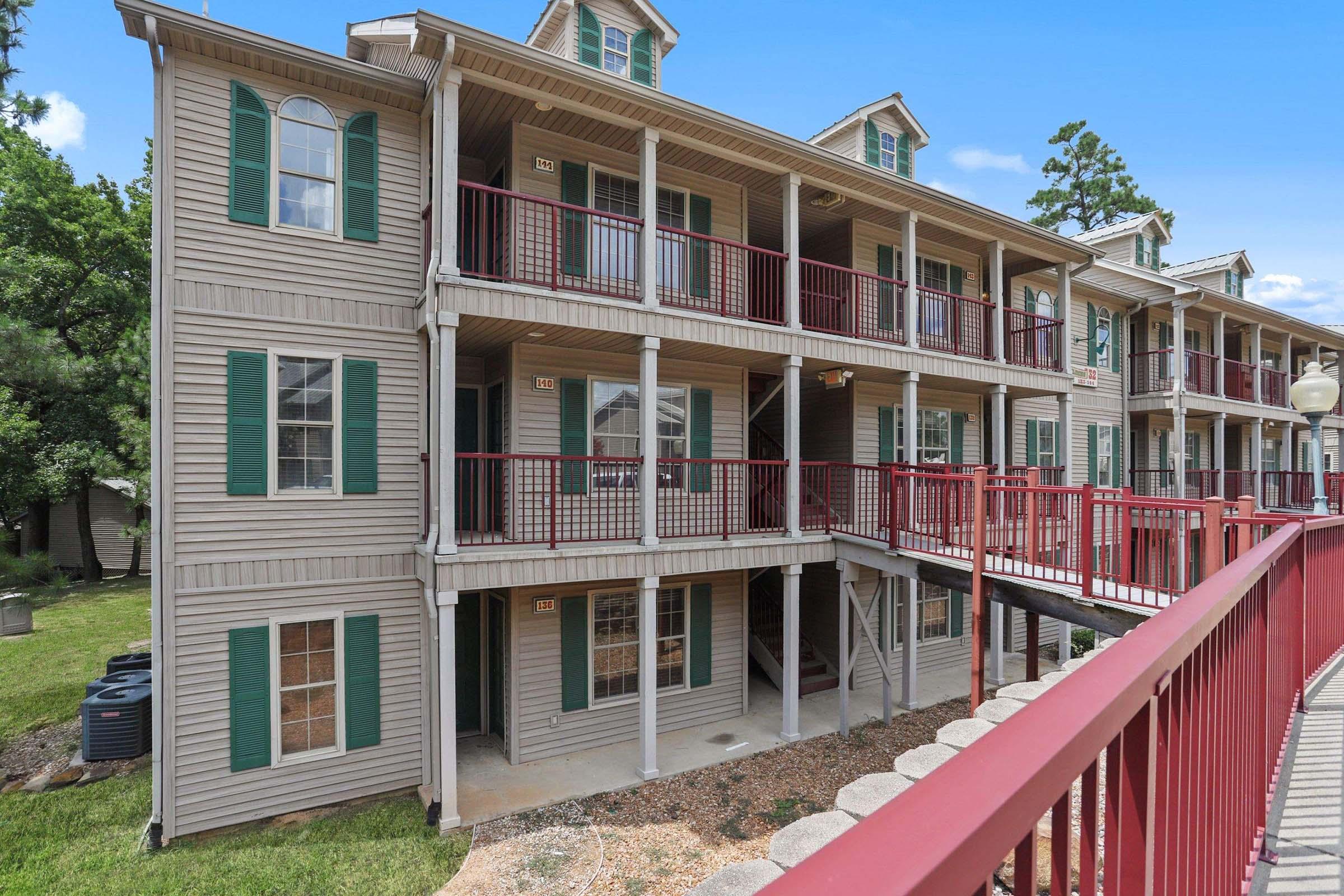 A multi-story apartment building with wooden exteriors, green shutters, and a red railing. The building features multiple balconies and staircases, surrounded by a grassy area.
