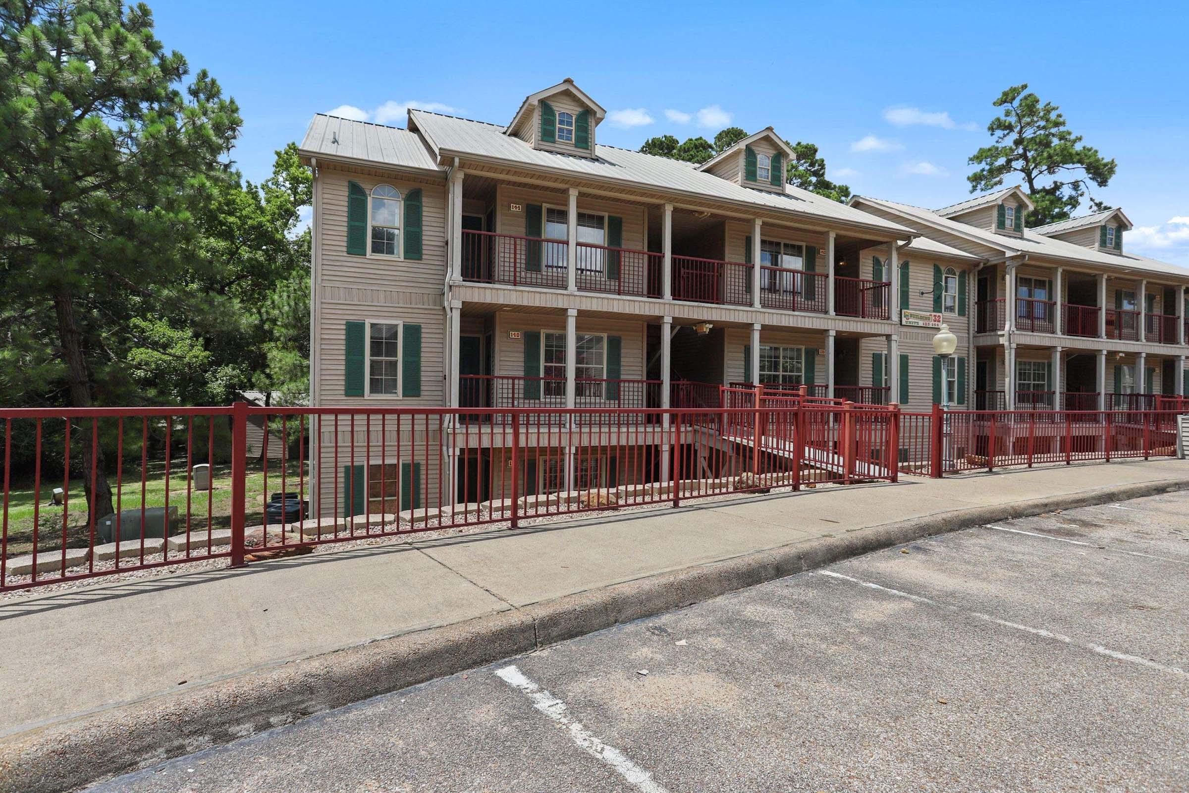 A multi-story building featuring a series of balconies with green shutters. The exterior is light-colored wood, and a red railing lines the walkway in front. The setting includes trees in the background and a paved area for parking.