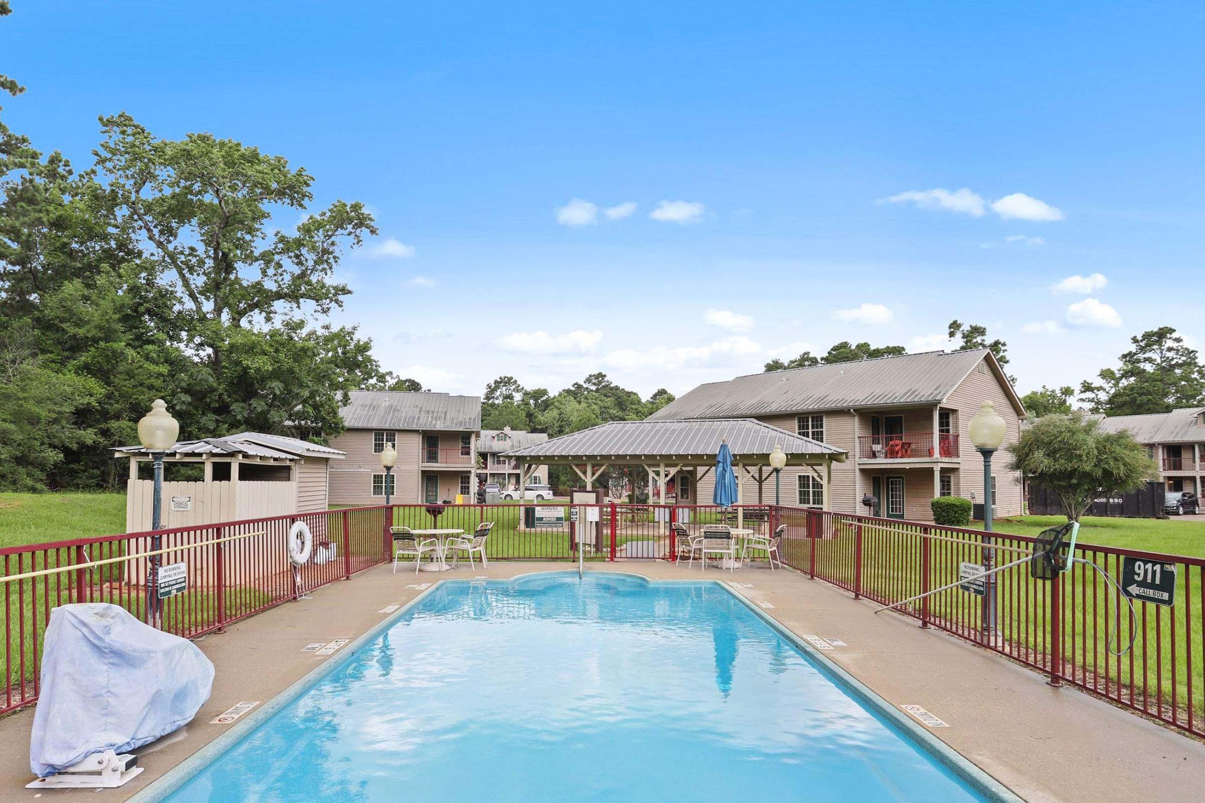 An outdoor swimming pool surrounded by a red safety fence, with several lounge chairs around it. In the background, there are two apartment buildings with balconies and a grassy area. A small gazebo is also visible nearby, and the sky is clear with a few clouds.
