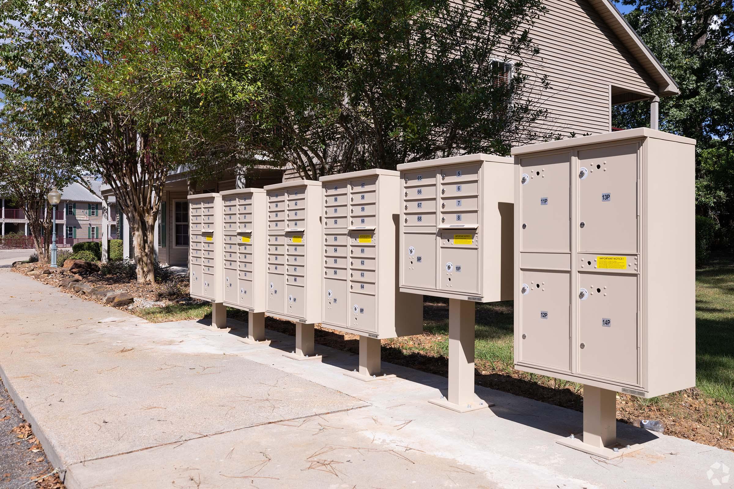 A row of beige postal mailboxes lined up on a pavement next to a residential building. The mailboxes have multiple compartments for mail delivery, with some featuring locks. Trees and grass are visible in the surrounding area, suggesting a suburban setting.