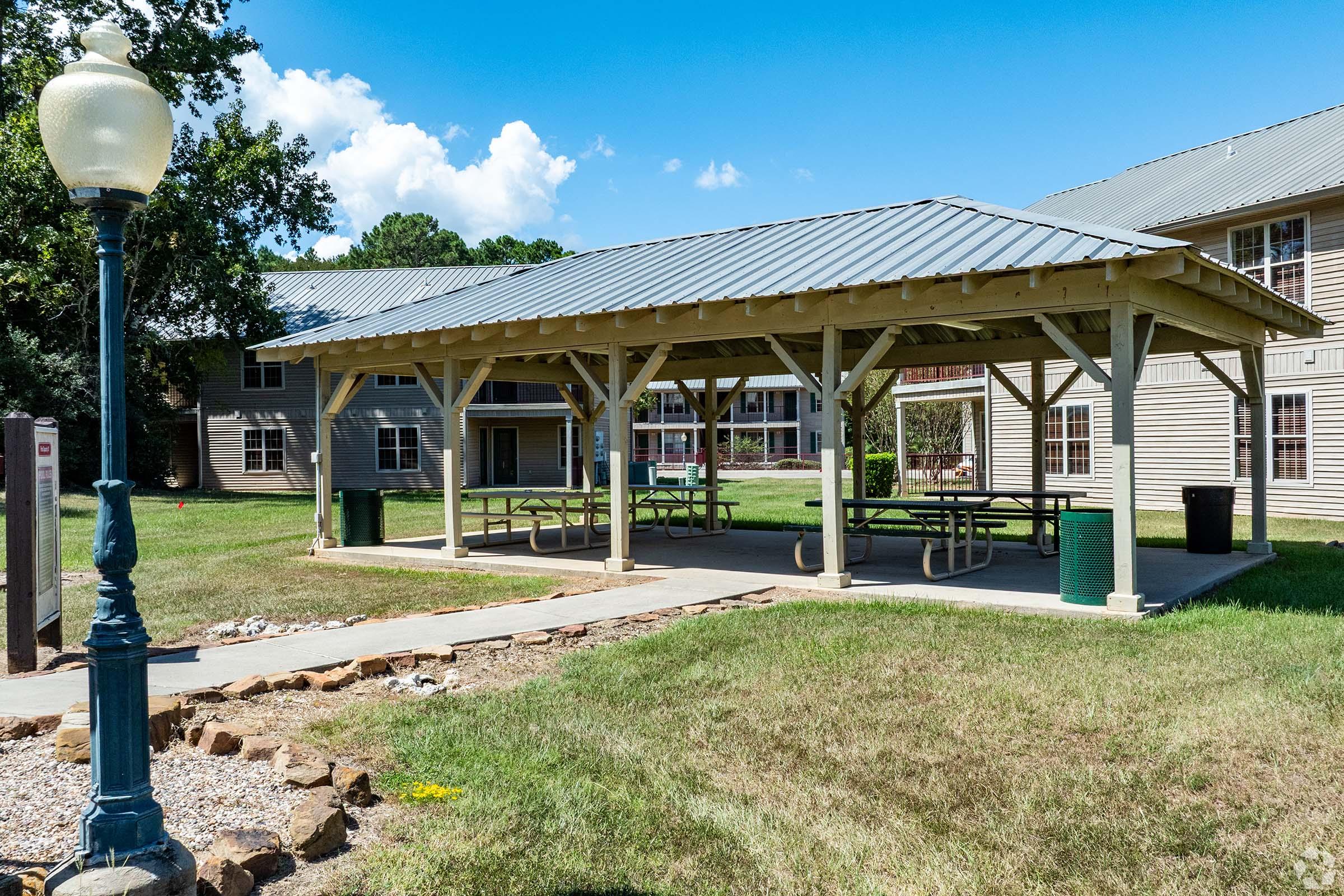 A covered outdoor pavilion with a wooden frame and metal roof, surrounding picnic tables. Green trash cans are located nearby, and a lamp post stands in the foreground. Surrounding the pavilion are grassy areas and a few buildings in the background under a blue sky with scattered clouds.