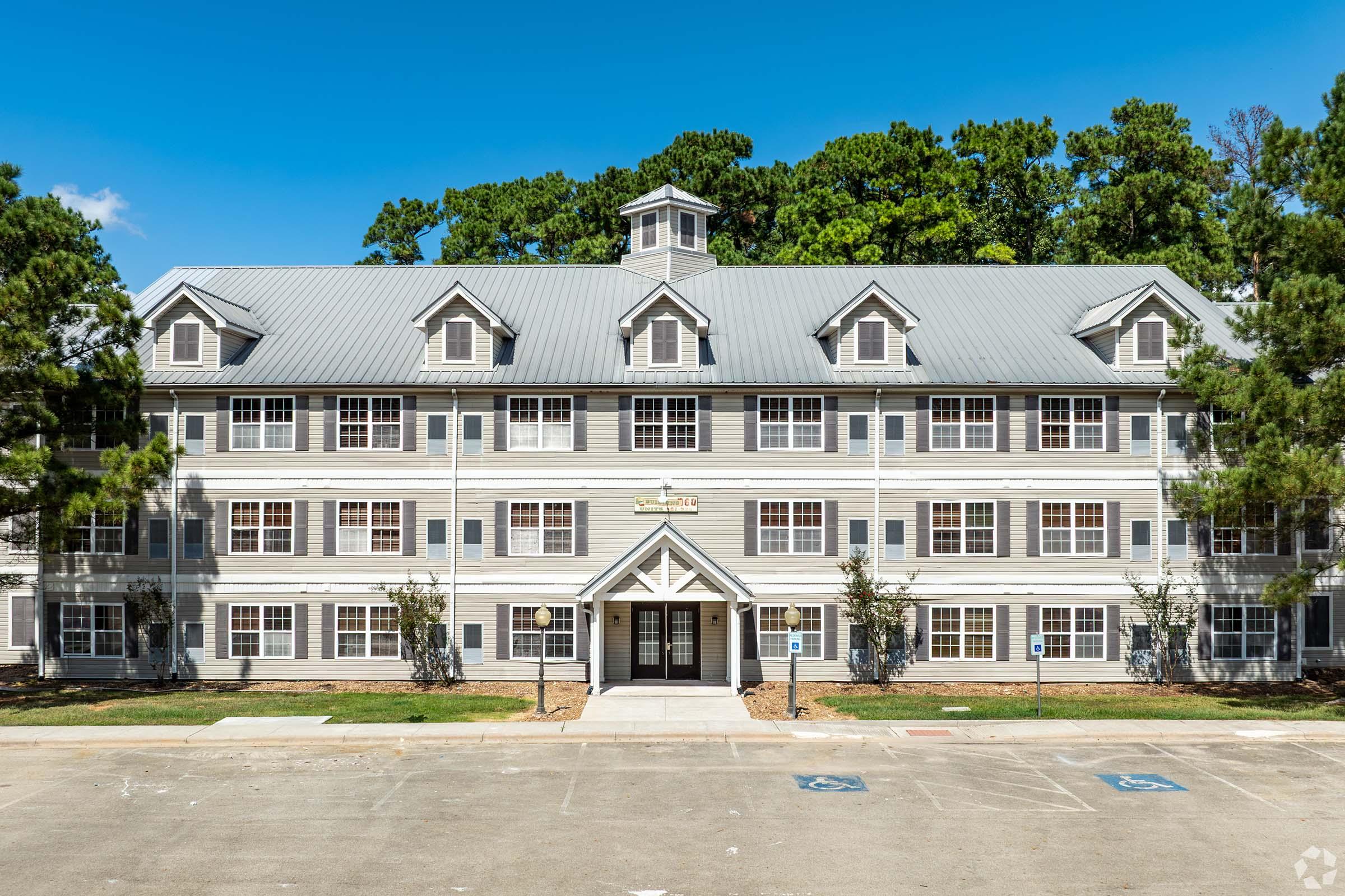 Three-story residential building with a grey exterior and white accents, featuring a central entrance with double doors and a peaked roof. Surrounded by landscaped trees, the structure has multiple windows and a wide driveway in front, with designated parking spaces visible.
