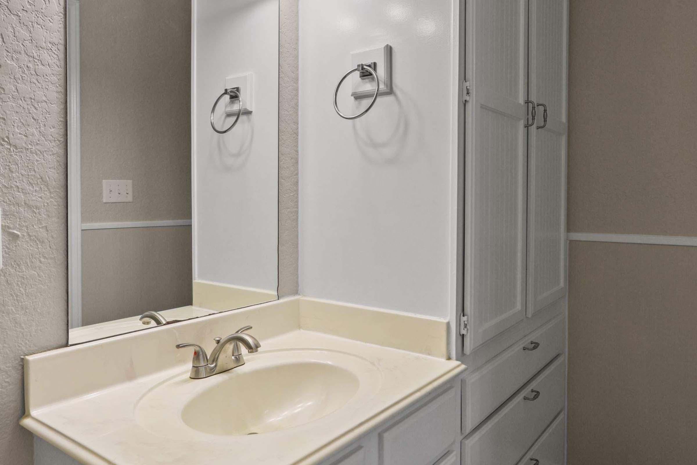 A clean bathroom with a white countertop and sink. There are mirrored cabinets above the sink, featuring a towel ring on the side. The walls are light-colored, and the overall decor is simple and modern.