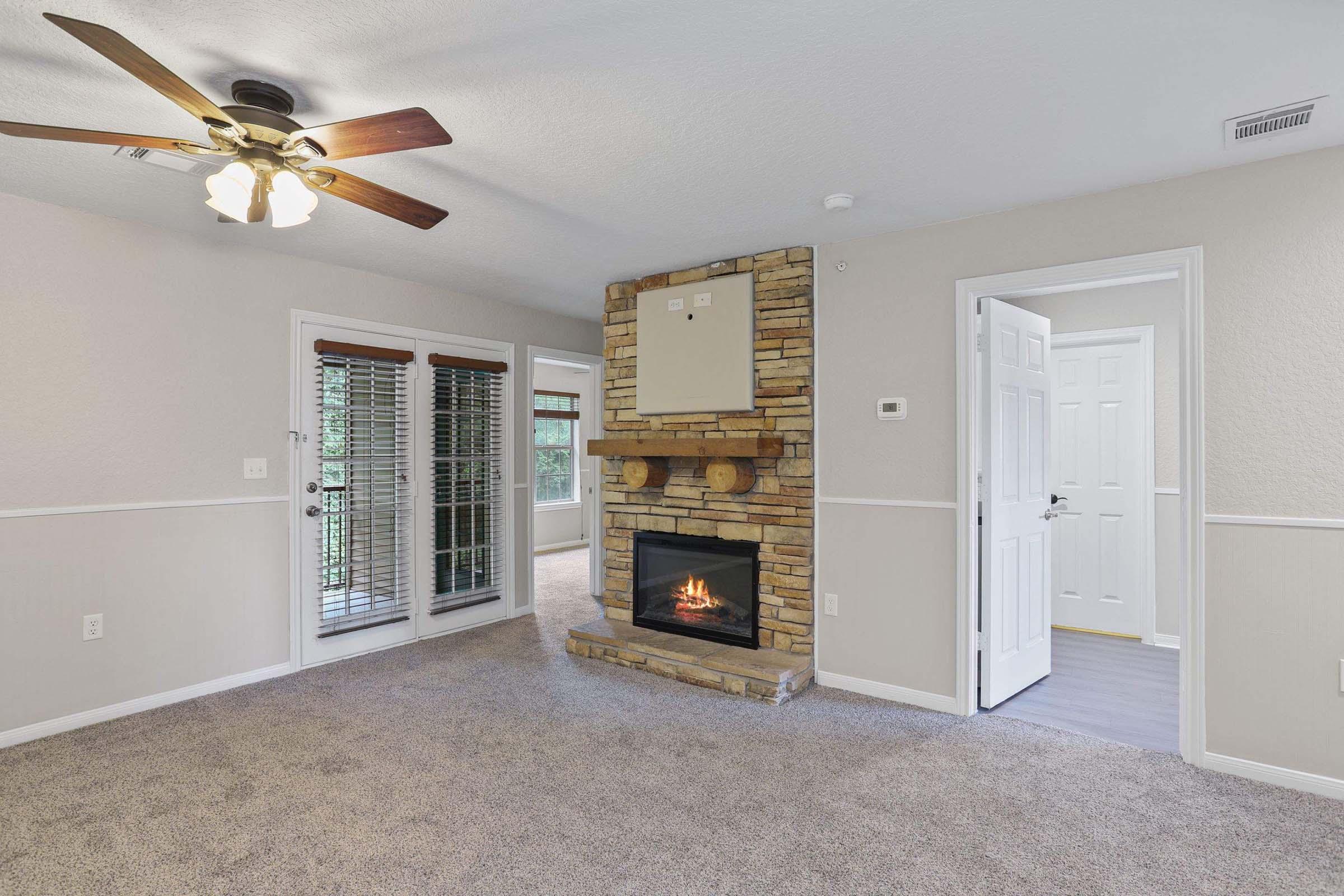 Cozy living room featuring a stone fireplace as the focal point, a ceiling fan, and large windows. The space has warm, neutral tones with plush carpeting and a modern design. A door leads to an adjacent room, while sliding glass doors open to the outside, adding natural light and access to fresh air.