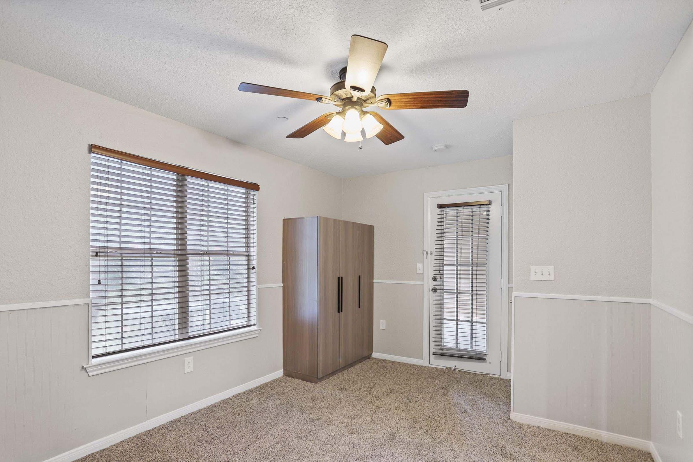 A small, empty room with beige carpet and light gray walls. A ceiling fan with wooden blades hangs above. There's a window with blinds on the left side, a closet or wardrobe in the corner, and a glass door leading outside on the right. Natural light fills the space, creating a bright atmosphere.