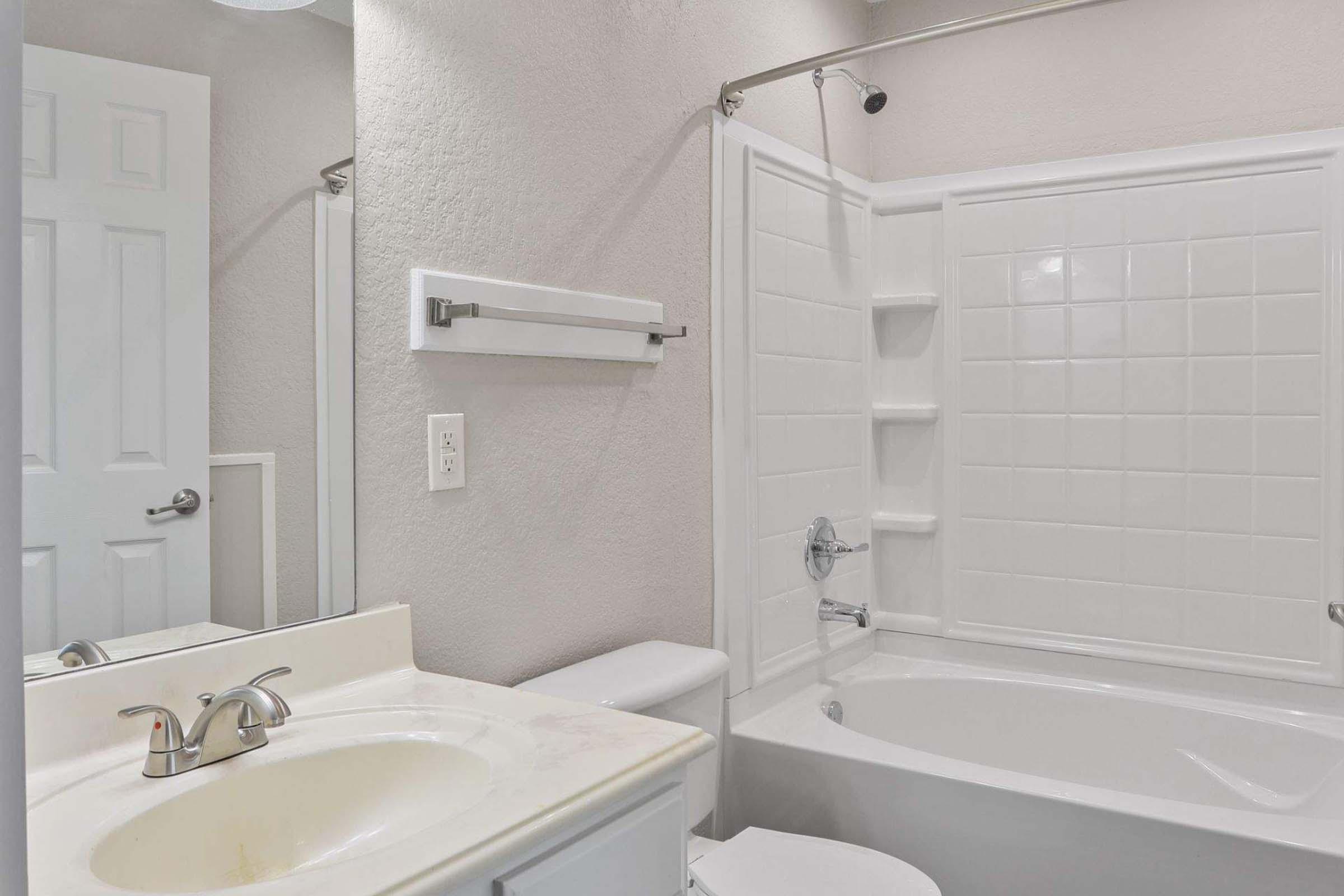 A clean bathroom featuring a bathtub with a shower, a white sink with a faucet, a wall mirror, and a towel rack. The walls are a light color, and there is a white door visible in the background. The room appears bright and well-maintained.