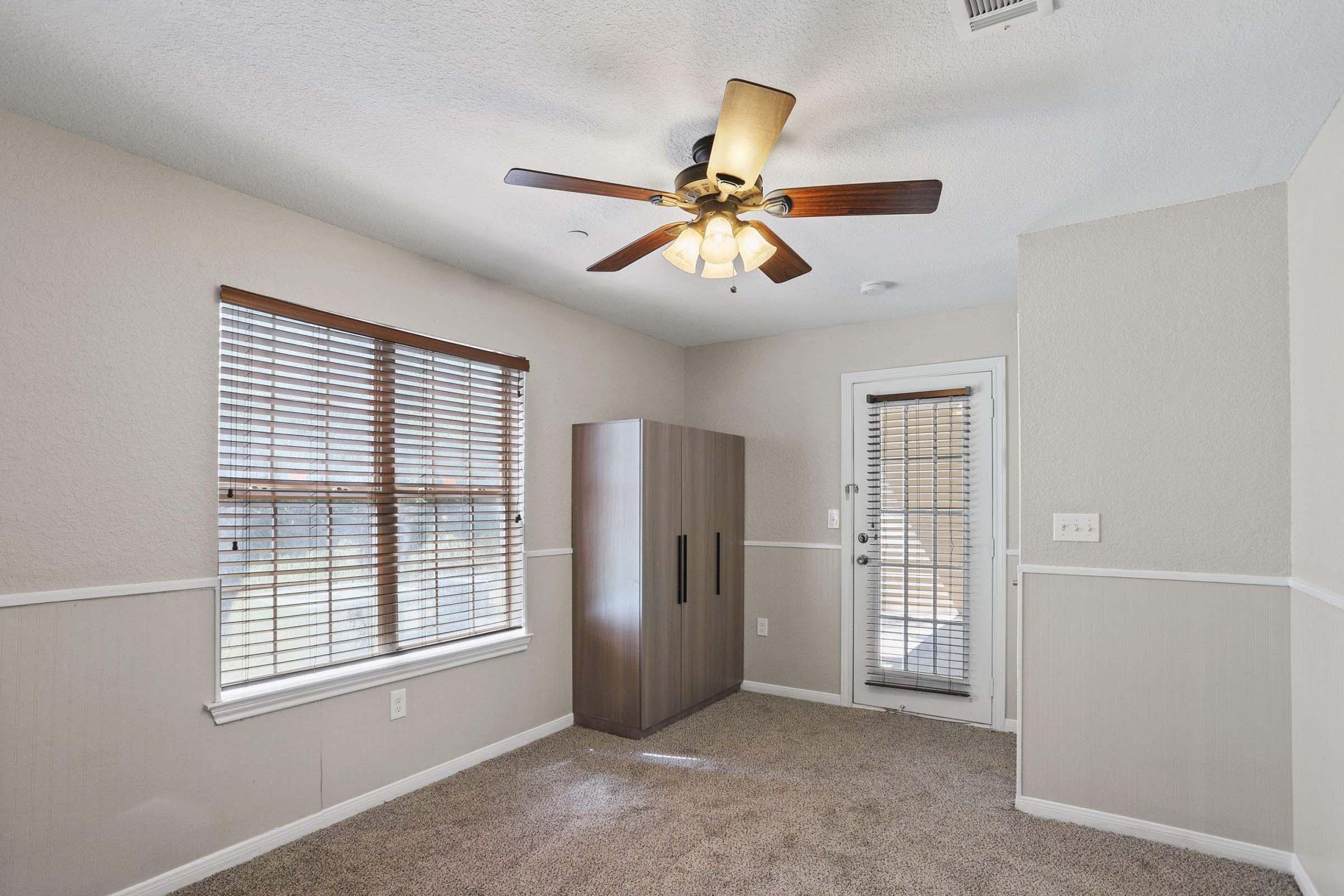 A small, well-lit room featuring a ceiling fan with lights, a window with blinds, and a door leading outside. There is a tall wardrobe against one wall and carpeted flooring, complemented by neutral-colored walls with wainscoting.