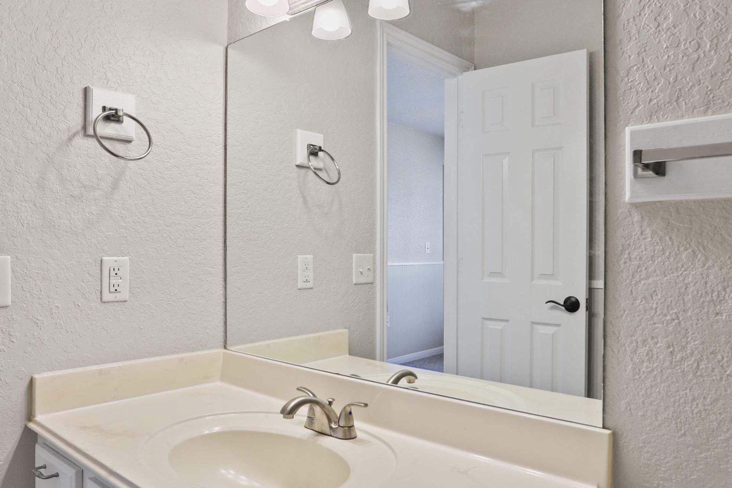 A bathroom featuring a light-colored countertop with a sink, a wall-mounted mirror, and modern fixtures. The room has light gray walls, a white door partially open, and a towel rack on the wall. Soft lighting illuminates the space, creating a clean and functional aesthetic.