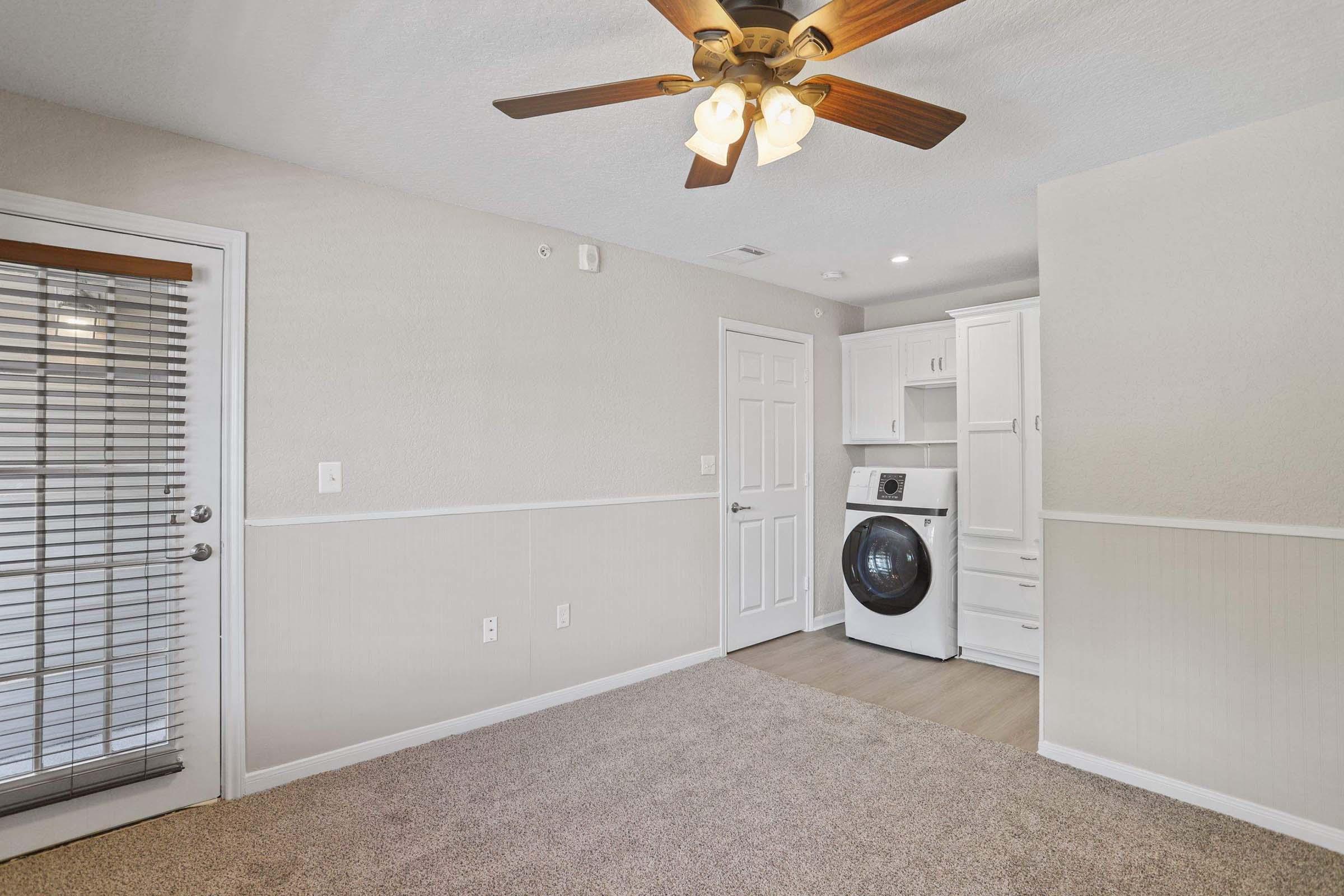 Interior view of a room featuring a ceiling fan with wooden blades, a laundry area with a washing machine, and beige walls. A door with blinds is visible, along with light-colored carpeting and a decorative wall panel. The room is well-lit and appears tidy and welcoming.