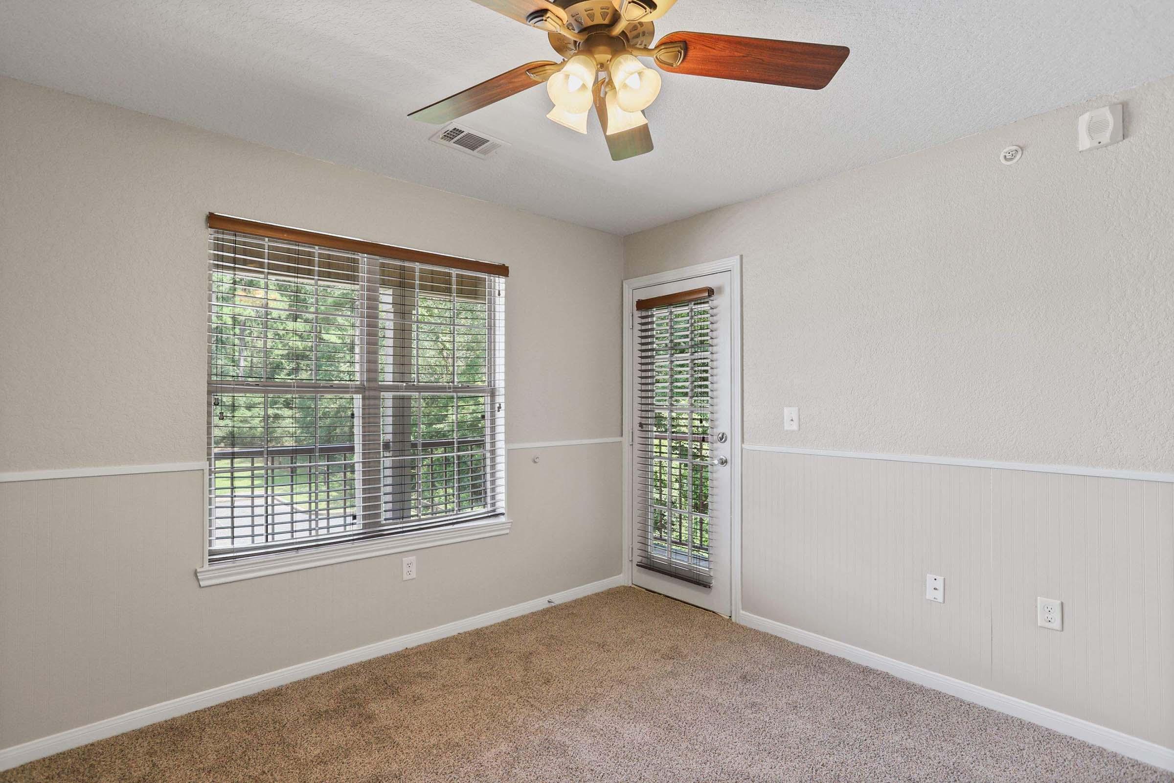 A sparsely furnished room featuring a ceiling fan with wooden blades, two windows with blinds, and a door leading outside. The walls are painted light beige, and the flooring is carpeted in a neutral tone. Natural light streams in, creating a bright and inviting atmosphere.