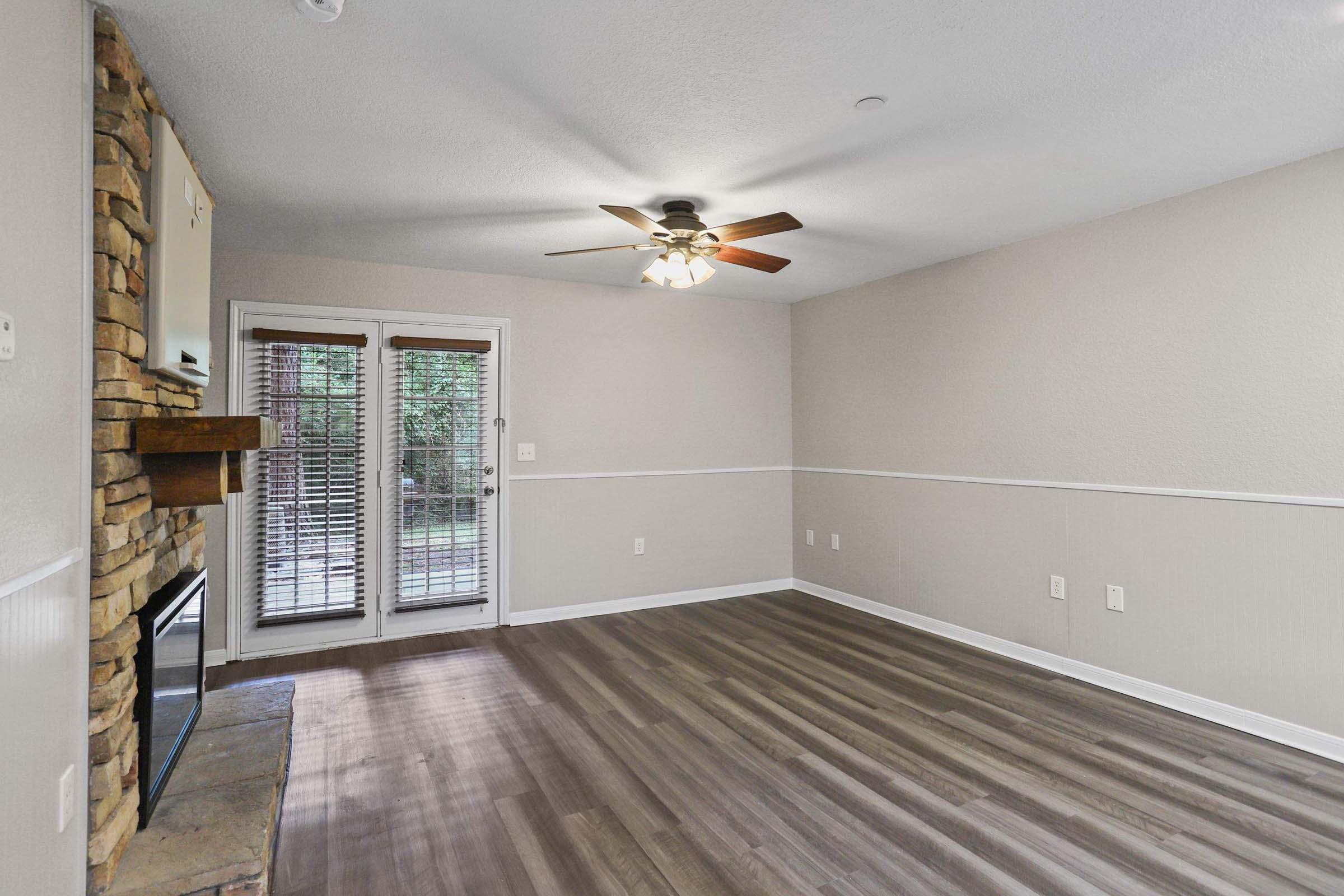 A spacious living room featuring light-colored walls, a ceiling fan, and a stone fireplace. The floor is adorned with wood-like laminate, and there are large windows with blinds allowing natural light. A set of double doors leads to an outdoor area.