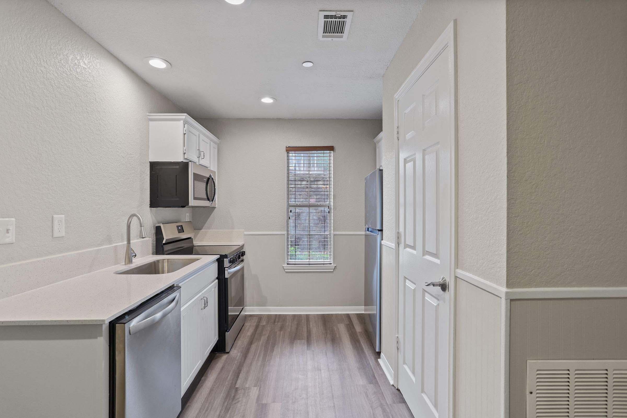A modern kitchen featuring white cabinetry, stainless steel appliances, and a light-colored countertop. The space includes a sink, microwave, and a window that lets in natural light. The flooring is a light wood look, and the walls are painted in neutral tones, creating a bright and inviting atmosphere.
