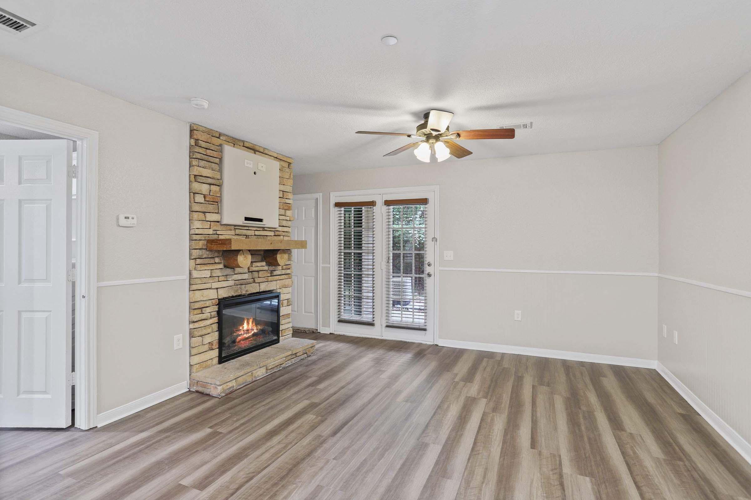 A cozy living room featuring a stone fireplace, wooden mantel, and ceiling fan. The room has light-colored walls and a laminate wood floor. A set of double doors leads to an outdoor area, and the space is well-lit with natural light.
