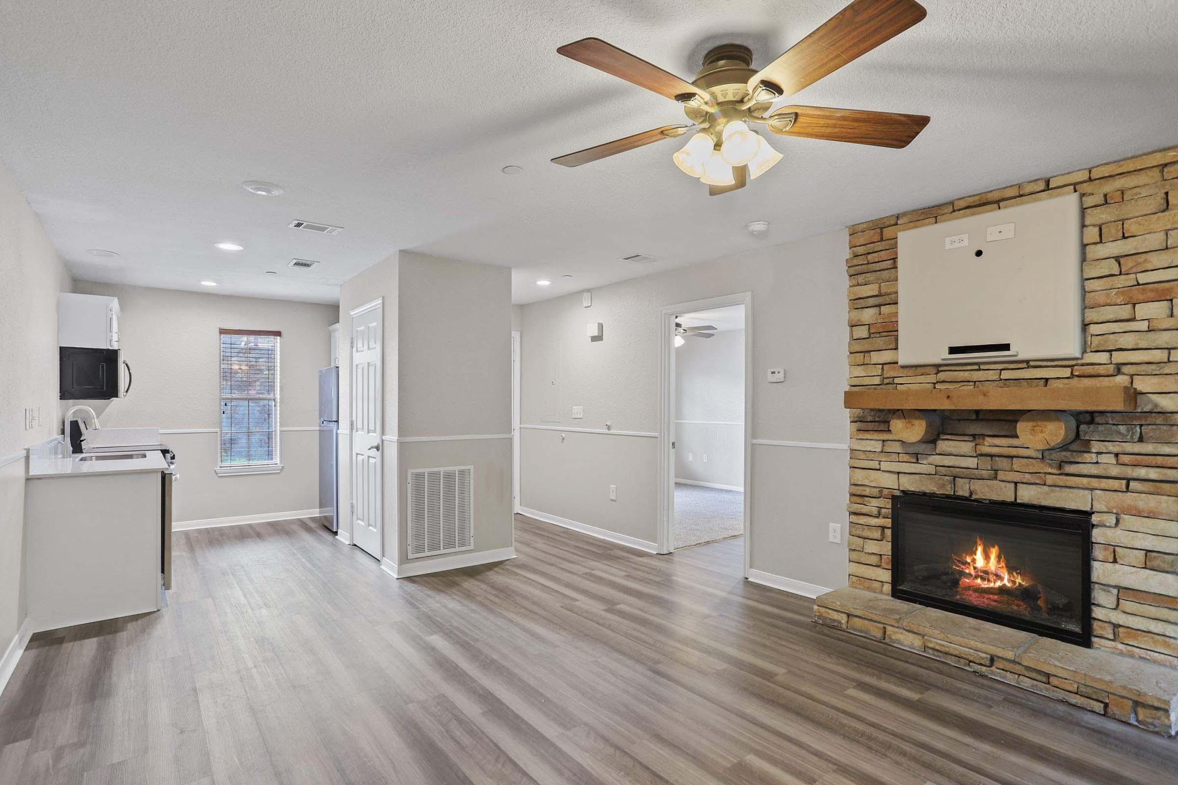 Interior of a modern apartment featuring a living area with a ceiling fan, hardwood-style flooring, a fireplace with a stone mantel, and a kitchen area with white cabinetry. Bright natural light comes through a window, and there is a door leading to another room. The space is well-lit and inviting.