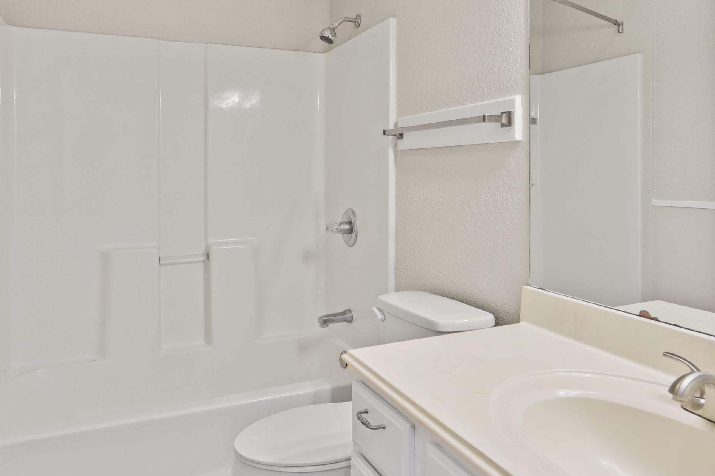 A clean and modern bathroom featuring a white bathtub with a shower, a toilet, and a vanity with a sink. The walls are light-colored, and there is a large mirror above the sink. The space is well-lit and organized, with minimal decor.