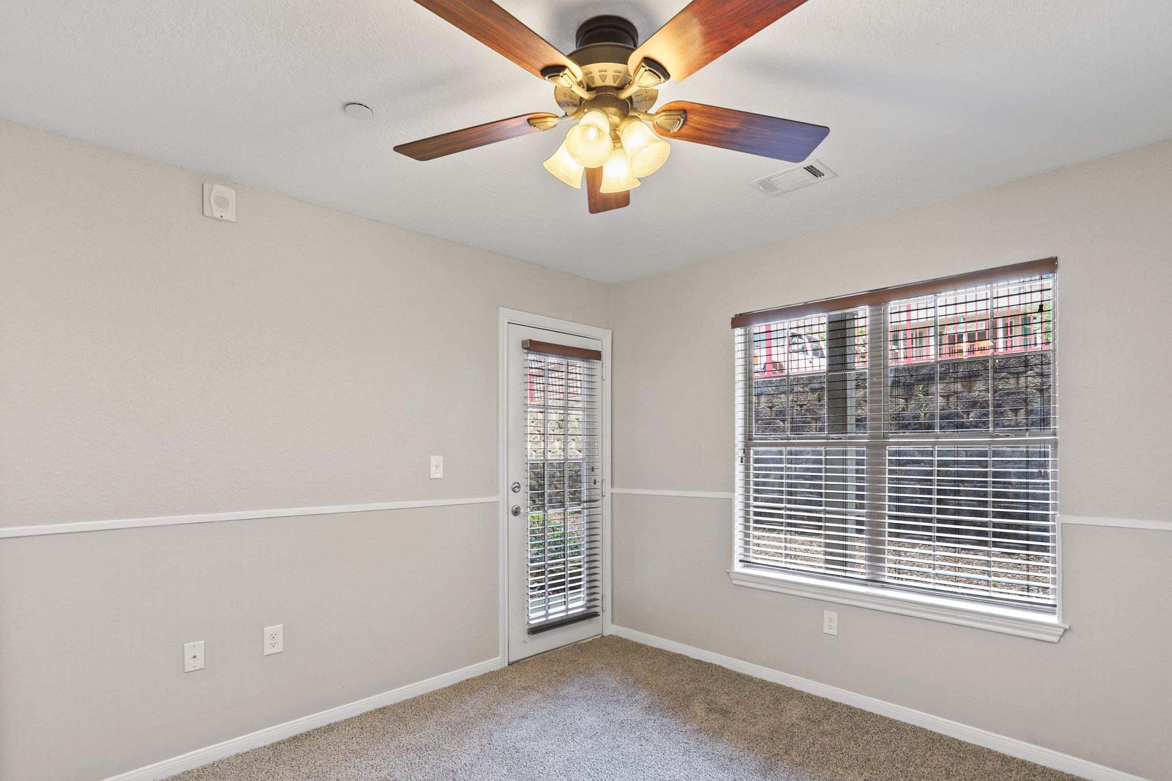 A well-lit room featuring beige walls and carpet. There is a ceiling fan with light fixtures, a glass door leading outside, and a window with blinds that lets in natural light. The room has a neat, inviting appearance, suitable for relaxation or work.