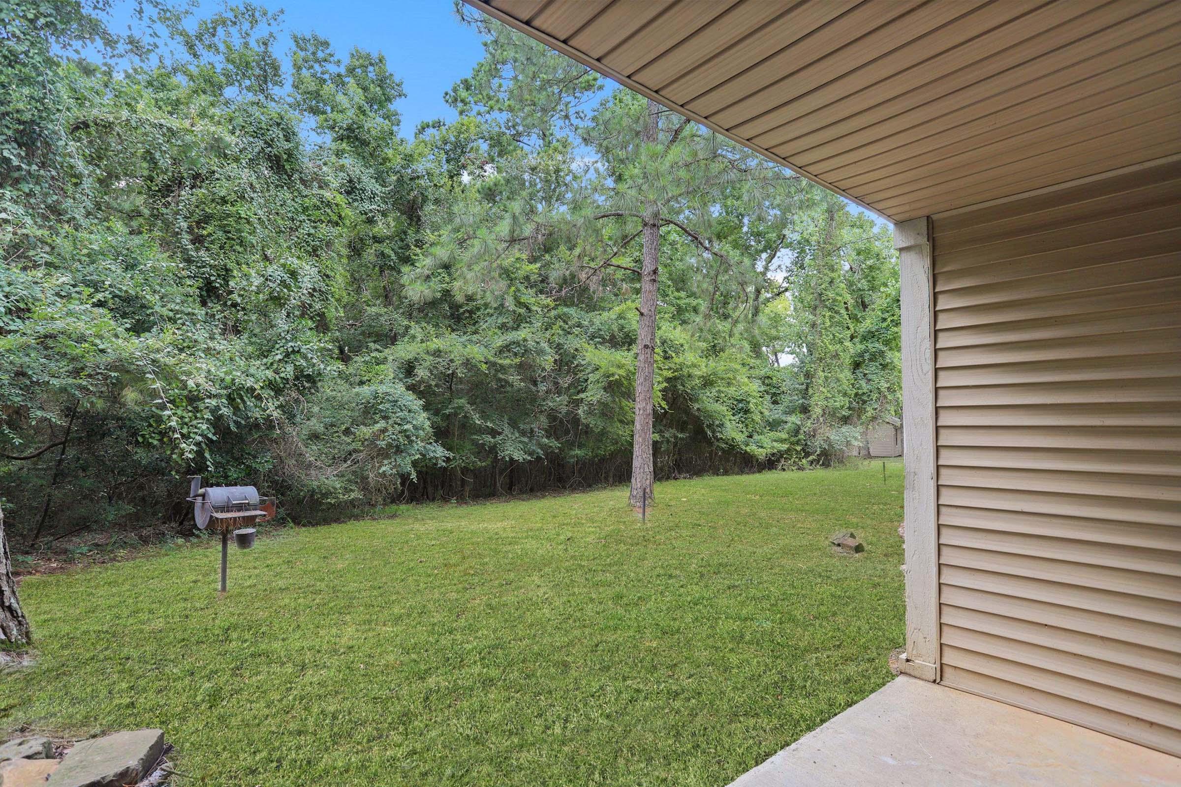 A view from a covered porch showing a lush green yard with tall trees and dense foliage in the background. There's a small BBQ grill to the left and a well-maintained lawn under bright, natural lighting. The scene conveys a peaceful, outdoor setting.