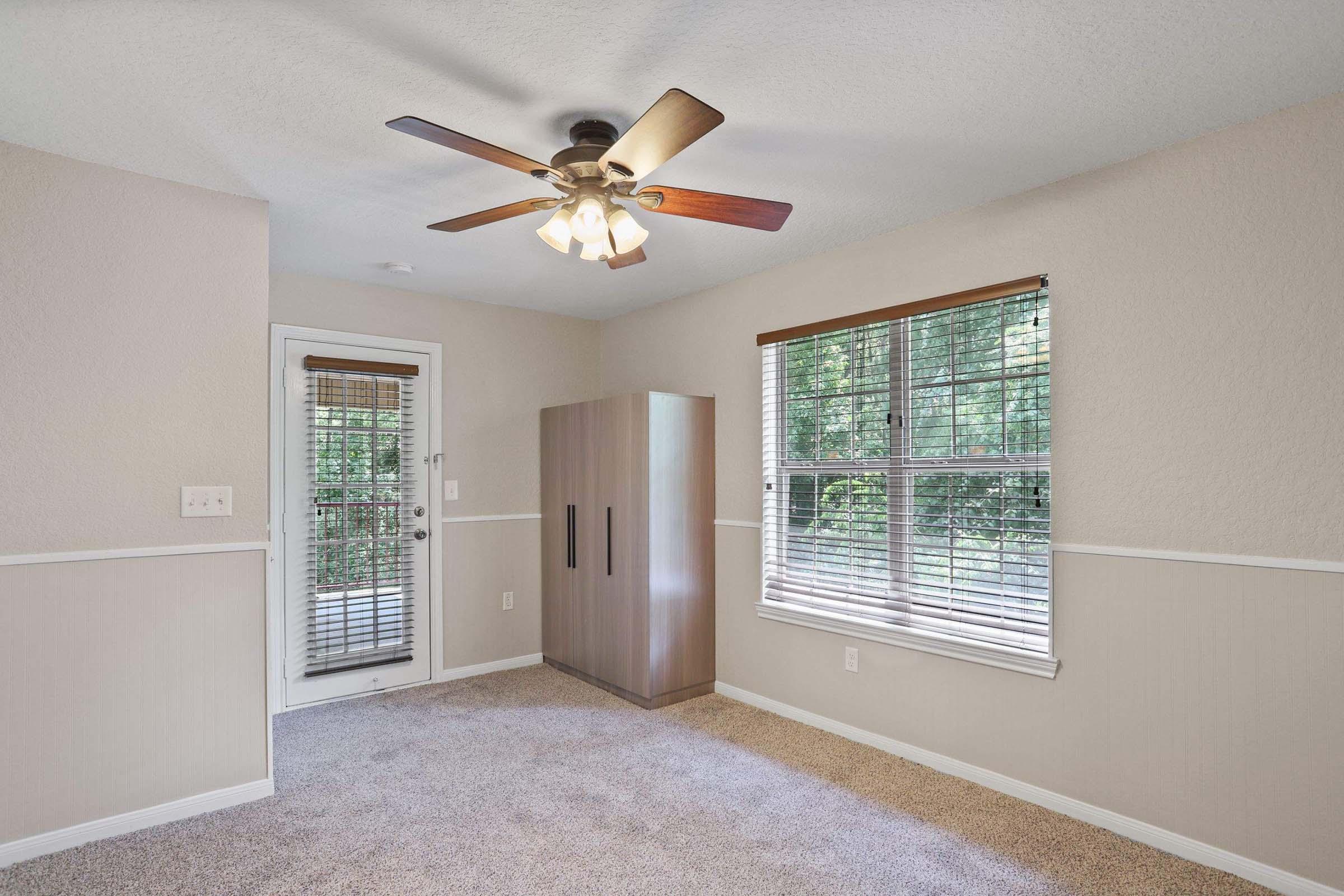 A cozy room featuring a ceiling fan, beige walls, and light brown carpet. There's a closed door leading outside and a window with blinds showing greenery outside. A modern wardrobe stands against the wall. Natural light fills the space, creating a warm and inviting atmosphere.