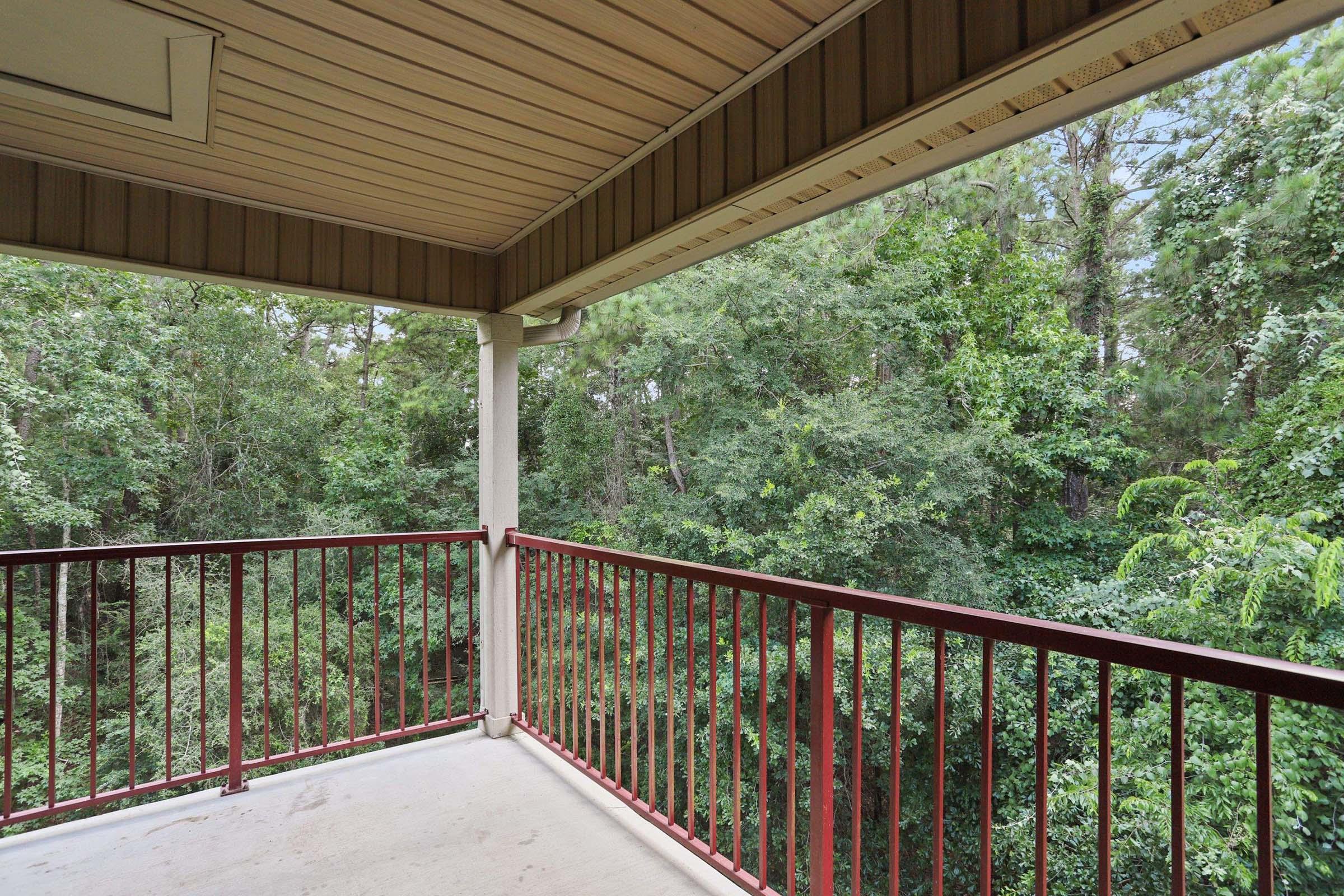 View from a balcony overlooking a lush green forest. The railing is red metal, and the flooring is concrete. The surrounding trees provide a dense, natural backdrop, creating a serene and peaceful atmosphere. The roof overhead adds shelter to the outdoor space.