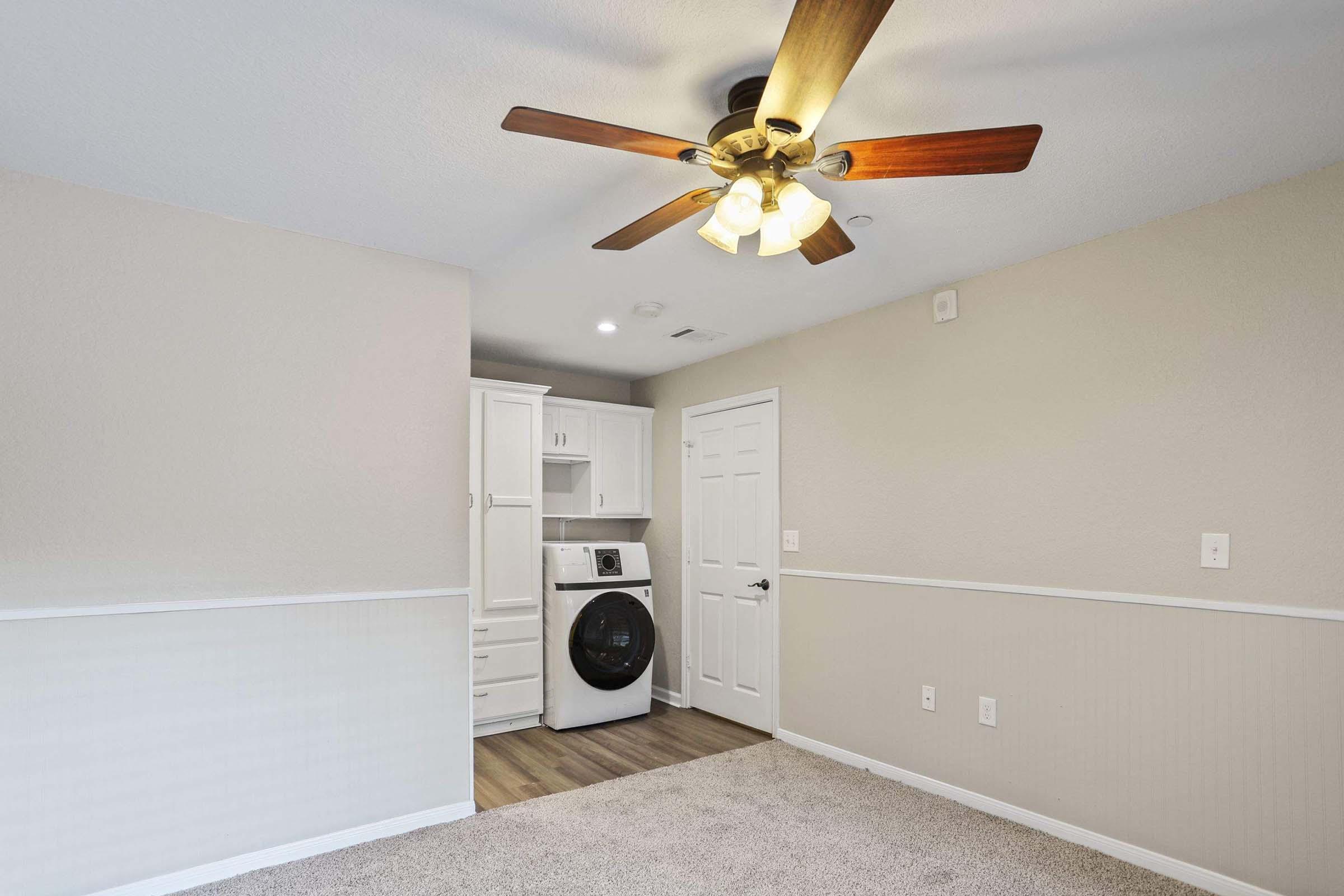 A well-lit laundry area featuring a ceiling fan, washer, and dryer. The room has beige walls with a white accent line and carpeted flooring. A door leads to another room, and there are built-in cabinets for storage, creating a functional and organized space.