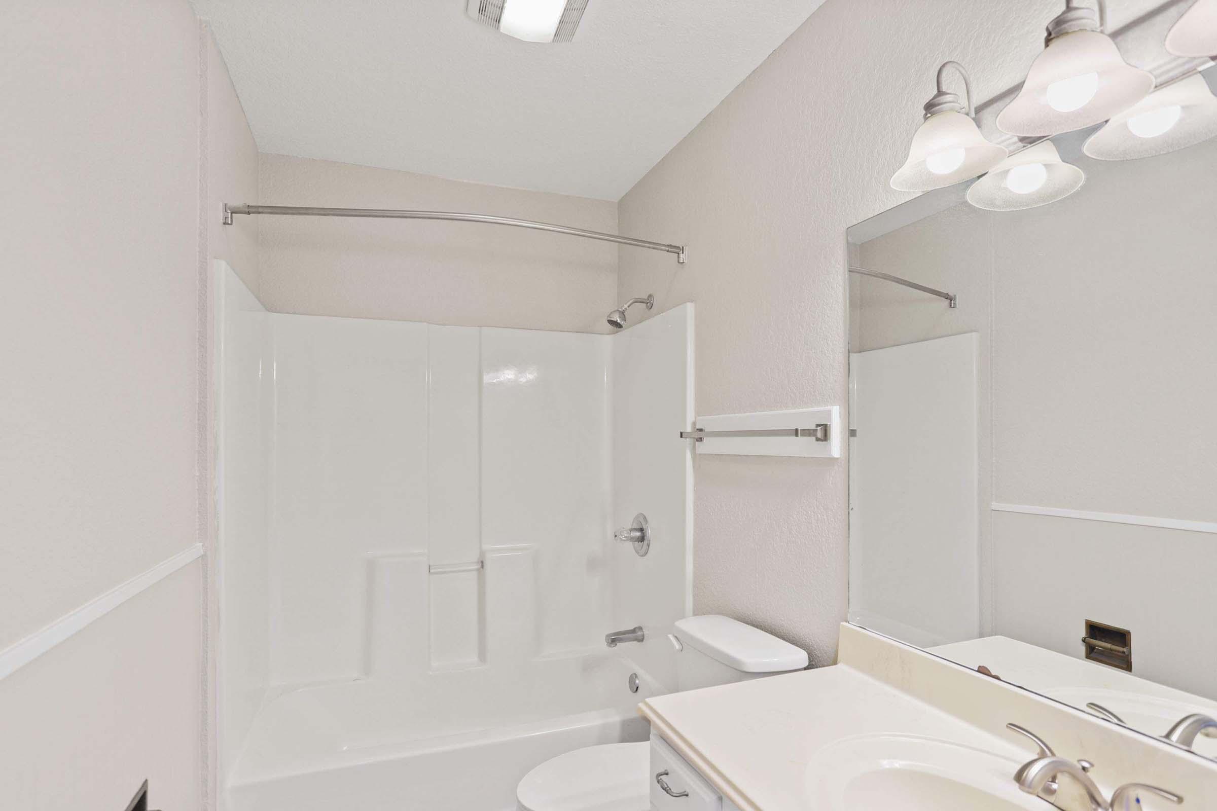 A clean and bright bathroom featuring a white bathtub with a shower, a toilet, and a vanity with a sink. The walls are painted a light color, complemented by a large mirror above the sink and three light fixtures above it. A towel rack is mounted on the wall beside the shower.