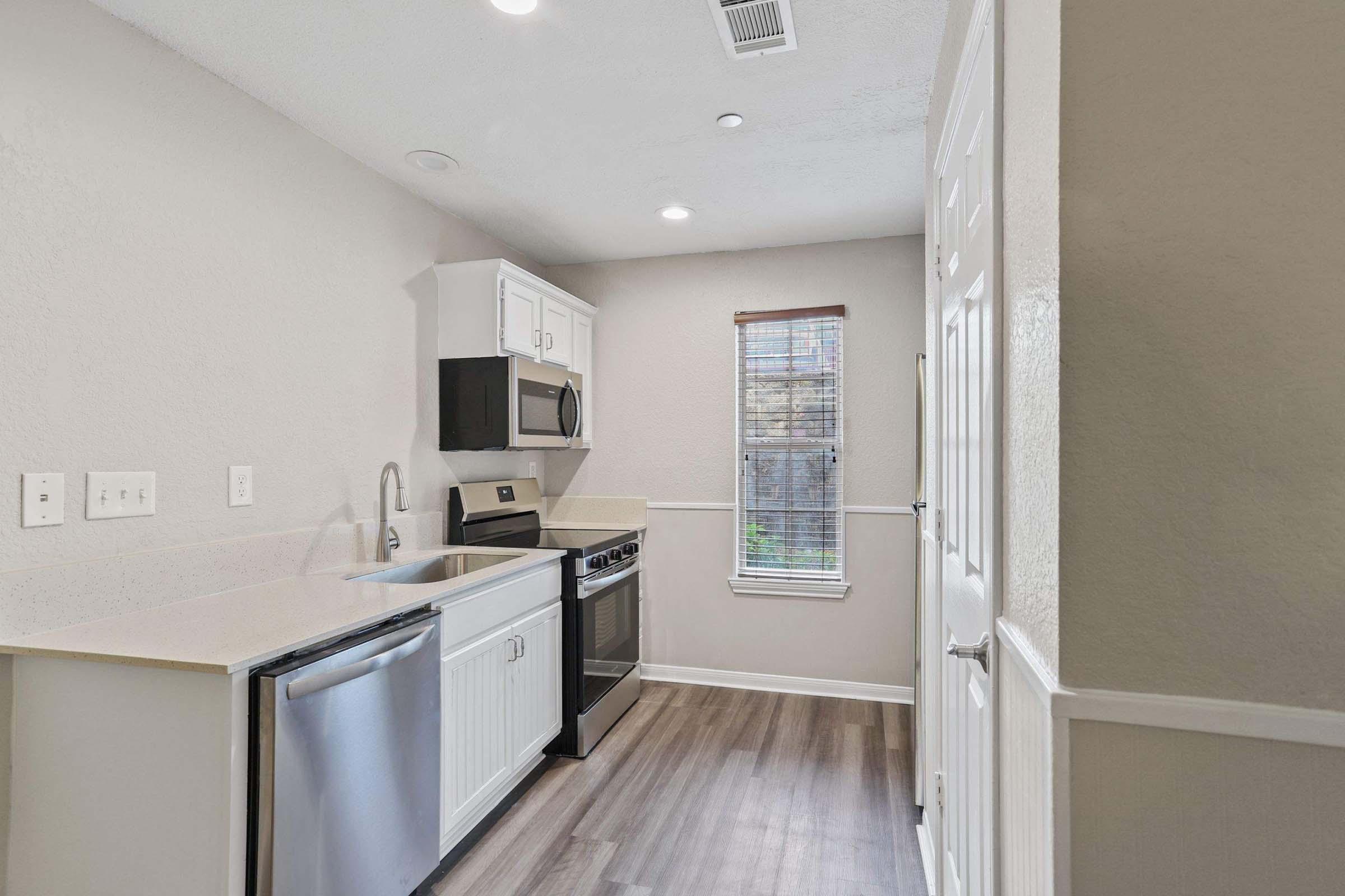 Modern kitchenette featuring white and black cabinetry, stainless steel appliances, including a microwave, oven, and dishwasher. Light-colored countertops with a sink are complemented by a window with blinds. The floor is made of wood-like laminate, creating a welcoming atmosphere.