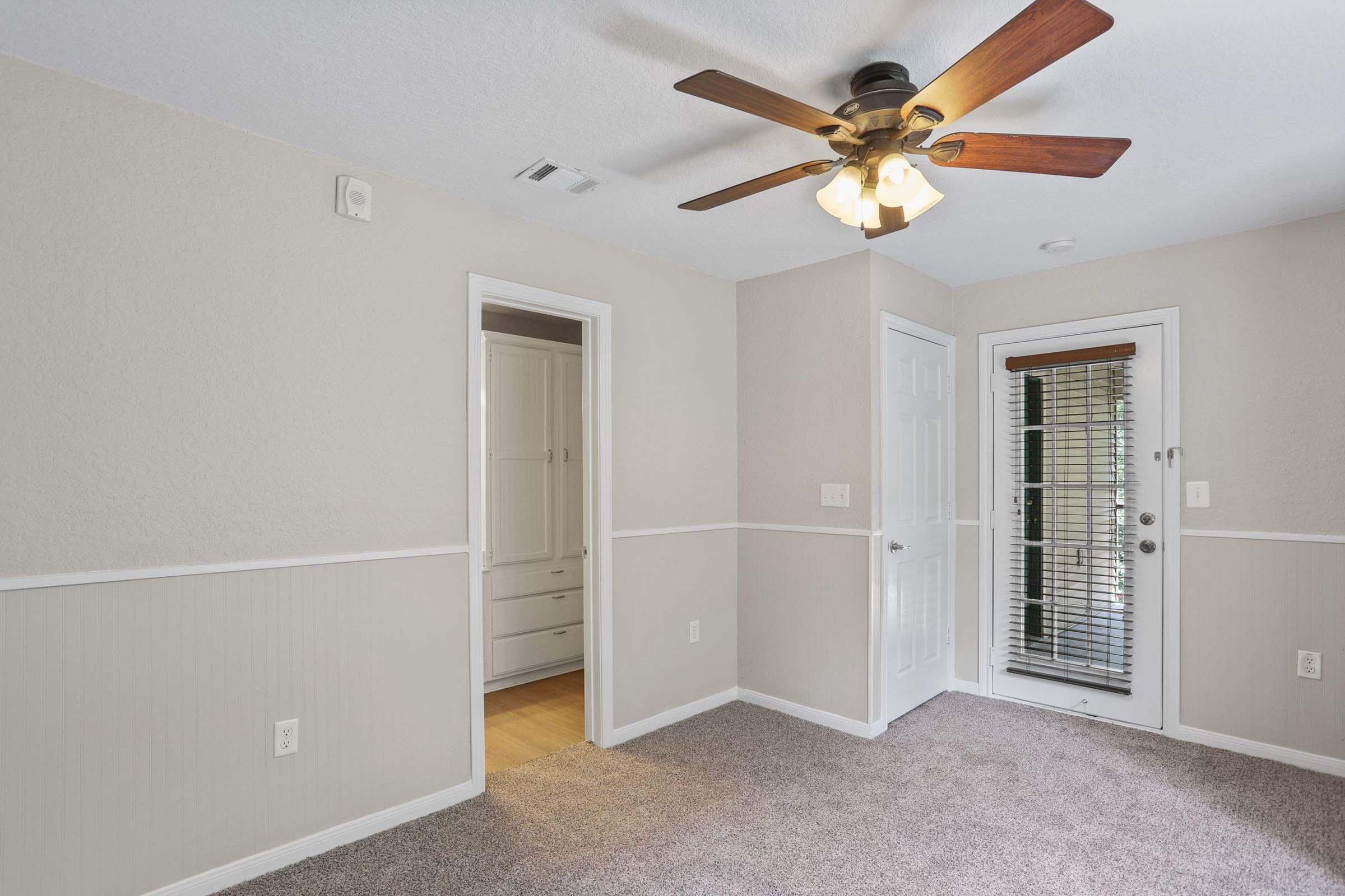 A cozy room featuring a ceiling fan with light, beige walls, and a carpeted floor. There is a door leading outside and another door that opens to a closet space. The room has natural light coming from the windows, creating a warm and inviting atmosphere.