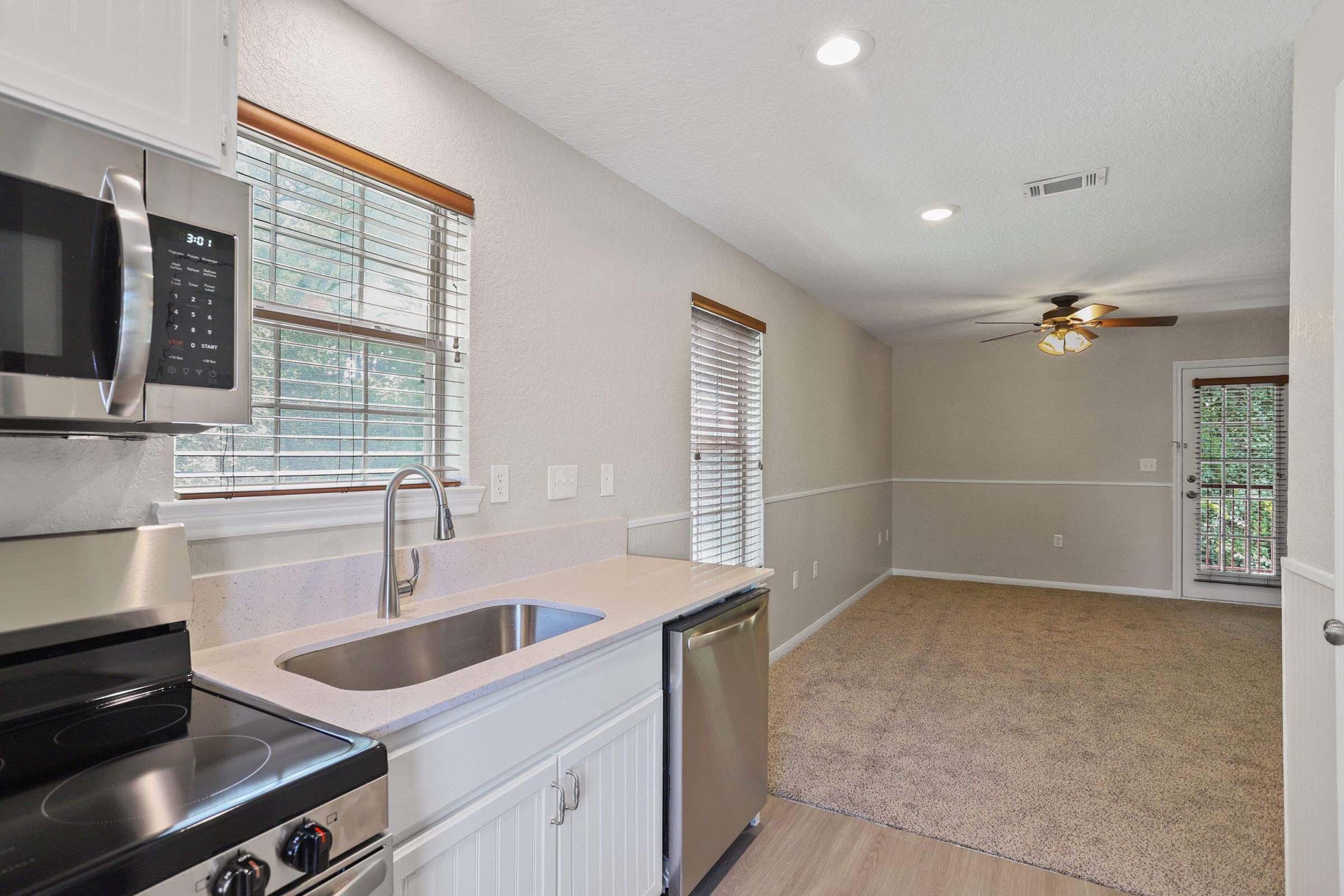 A modern kitchen featuring stainless steel appliances, including a microwave and oven, paired with a sleek sink. The space has light-colored walls and wooden window frames, allowing natural light to brighten the area. Adjacent to the kitchen is a carpeted living room with a ceiling fan and sliding door access to the outdoors.