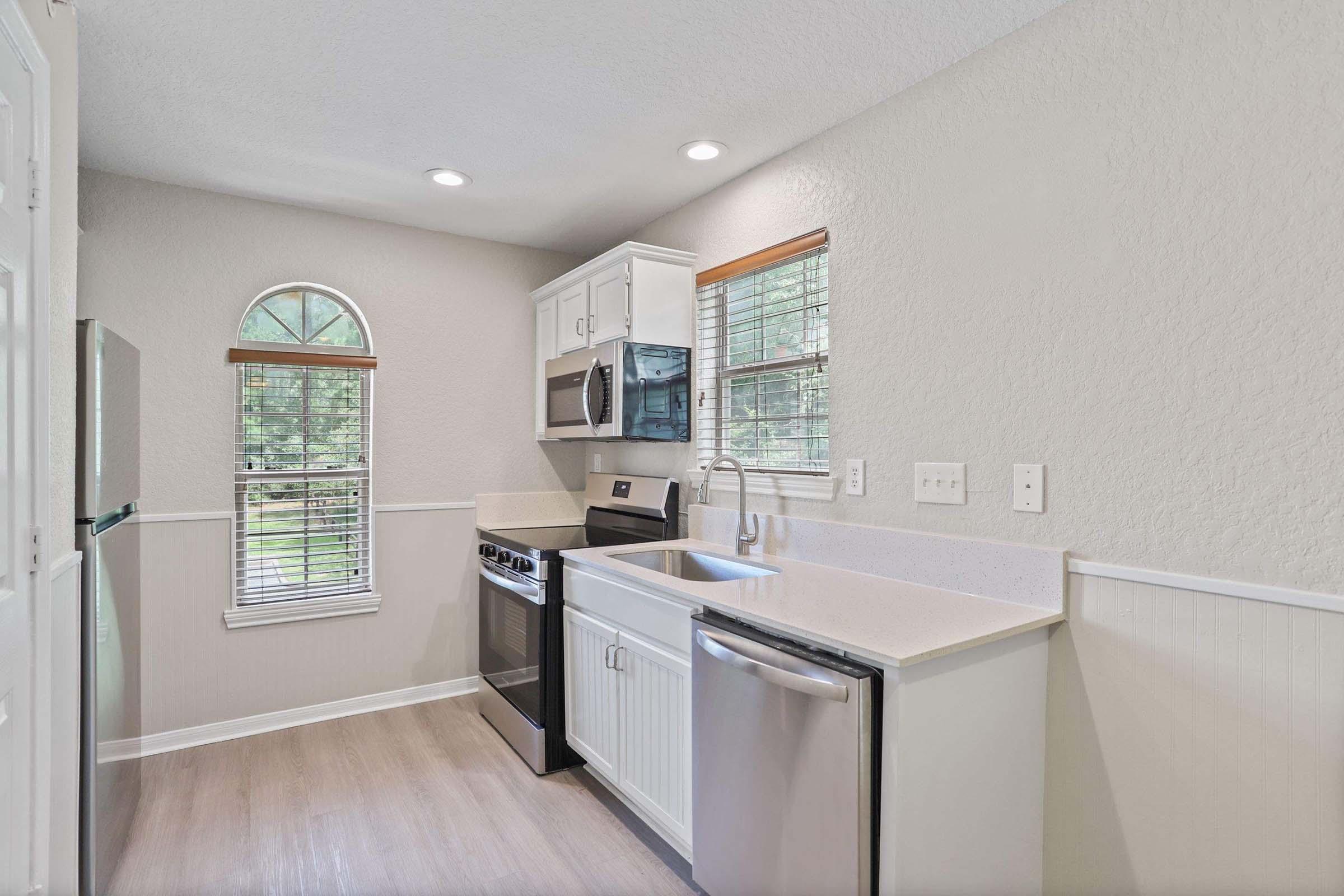 Modern kitchen with white cabinetry, quartz countertop, stainless steel appliances including a refrigerator, stove, and microwave. Features a window with natural light and a light gray wall color, creating a bright and welcoming atmosphere.
