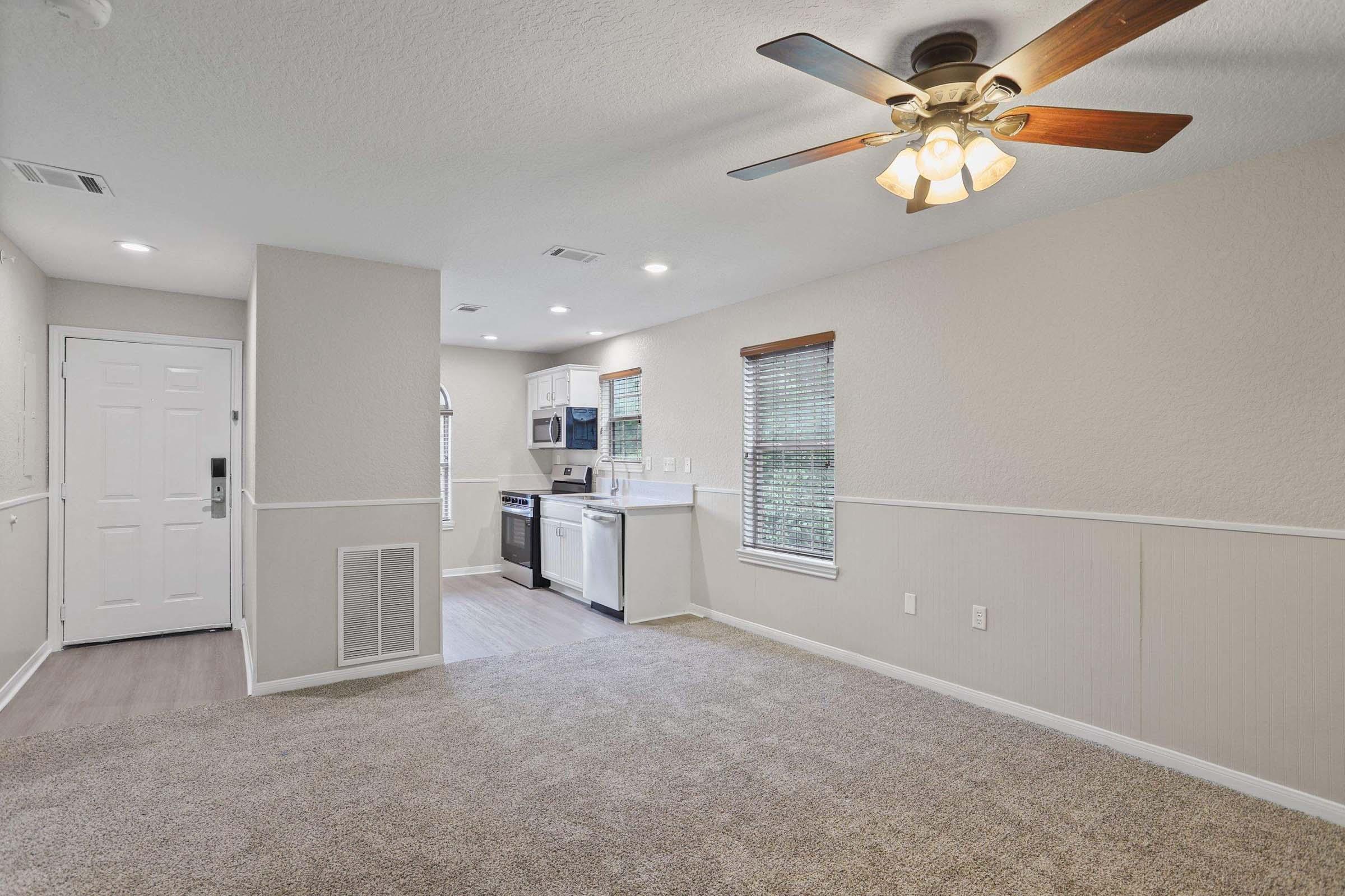 A well-lit interior of a modern living space featuring light-colored walls, a ceiling fan, and a cozy carpeted floor. To the left, there's a doorway leading outside, while the kitchen area is visible with white cabinets and appliances. Large windows with blinds allow natural light to enter the room.