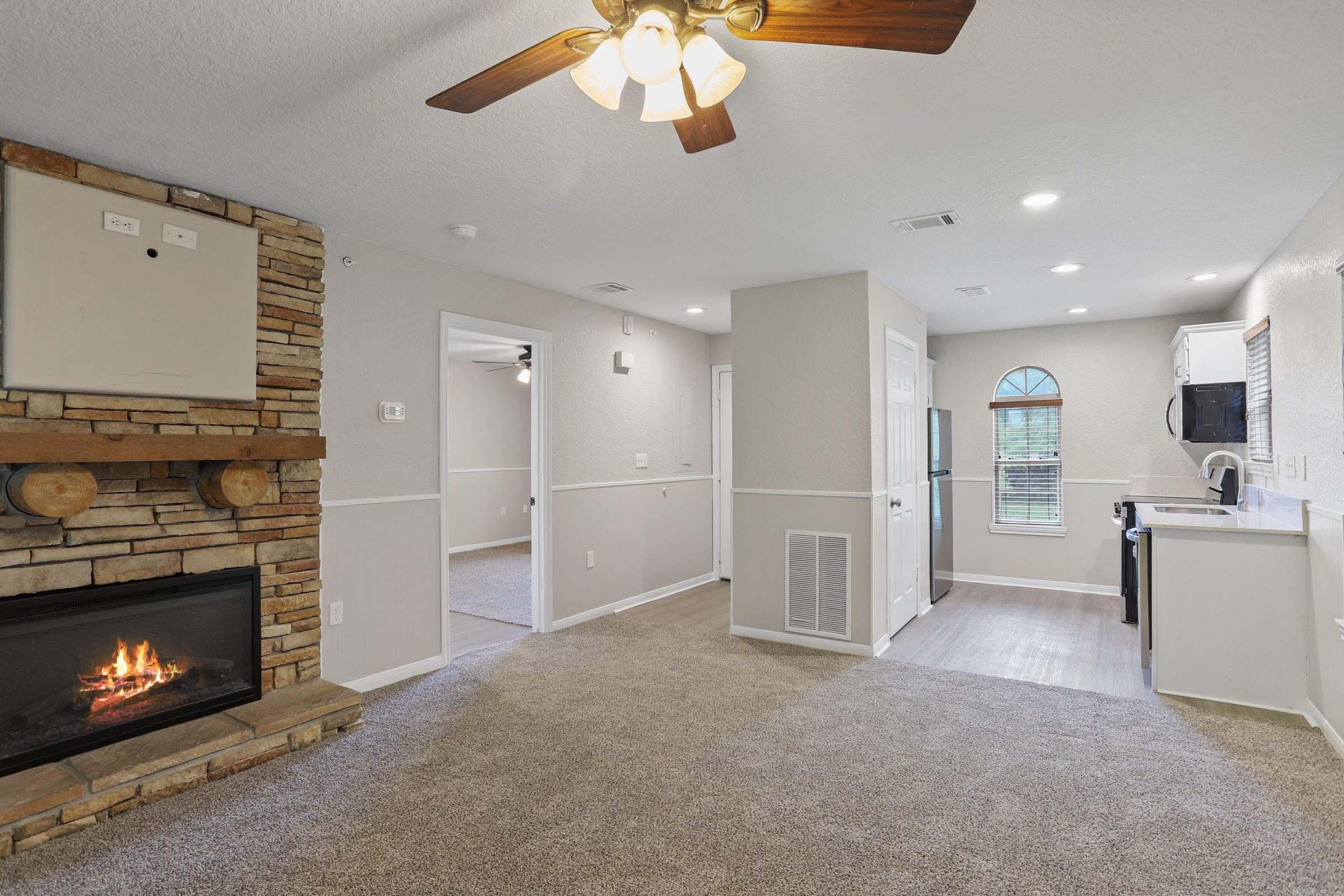 Cozy living space featuring a stone fireplace, ceiling fan, and carpeted flooring. The room is well-lit with natural light coming from a window. A doorway leads to a kitchen area, and the walls are painted in neutral tones, creating a warm and inviting atmosphere.