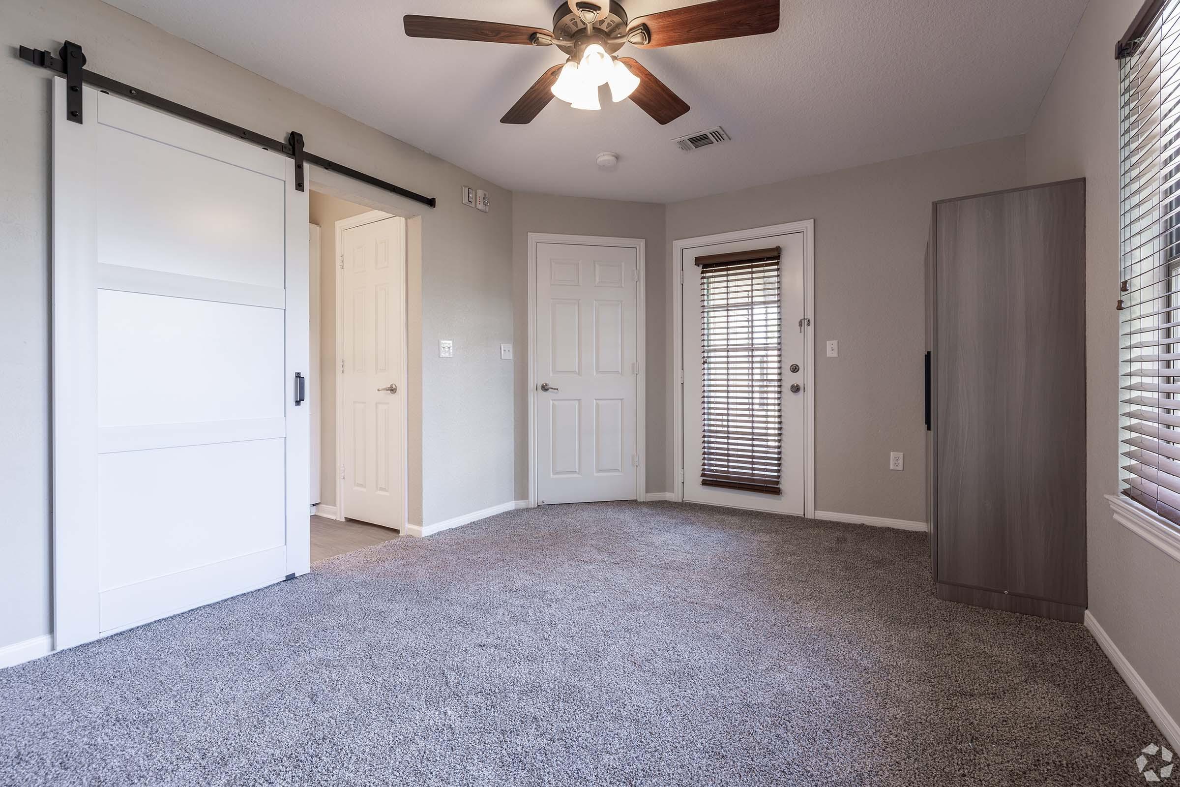 A well-lit, empty room featuring light gray walls and a carpeted floor. There's a ceiling fan, a sliding barn door, and two white doors leading to other areas. A window with blinds allows natural light in, and a small storage cabinet is positioned against one wall.